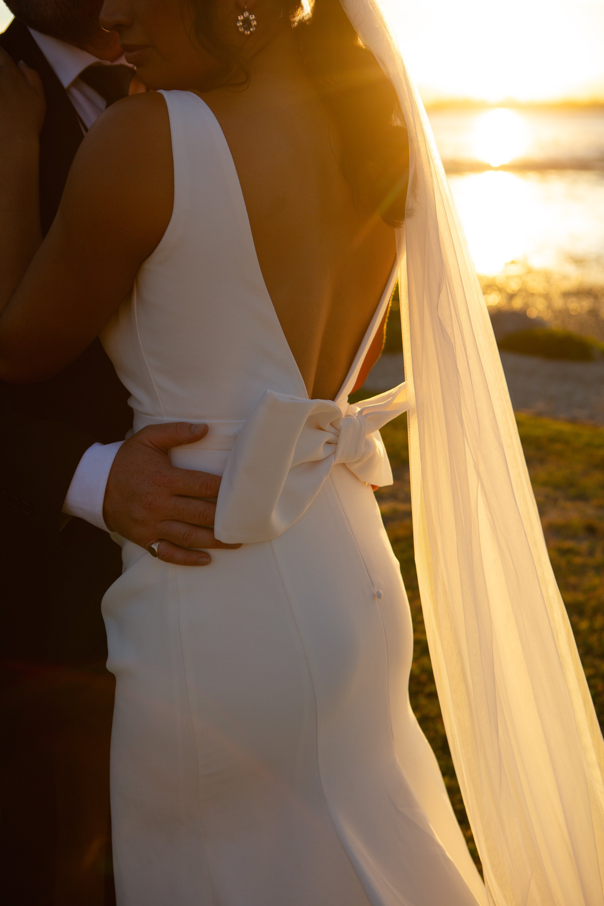A bride and groom embrace on a beach at sunset, with the bride wearing a white wedding dress with a bow at the back and a veil, and the groom wearing a dark suit.
