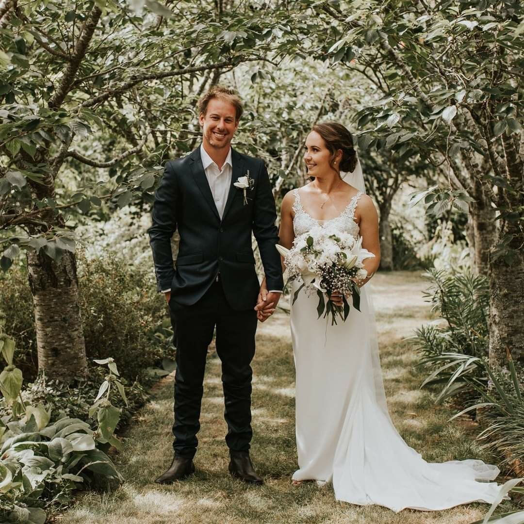 A bride and groom holding hands in a garden, with trees overhead, during a wedding ceremony.