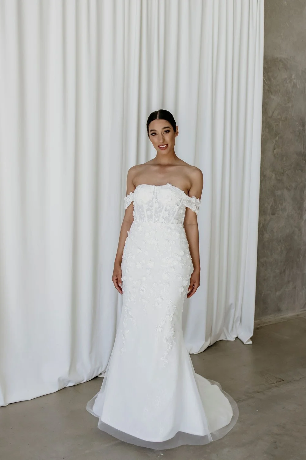 A woman in a white off-the-shoulder wedding gown, standing in front of white curtains with a marble wall on the side, smiling at the camera.
