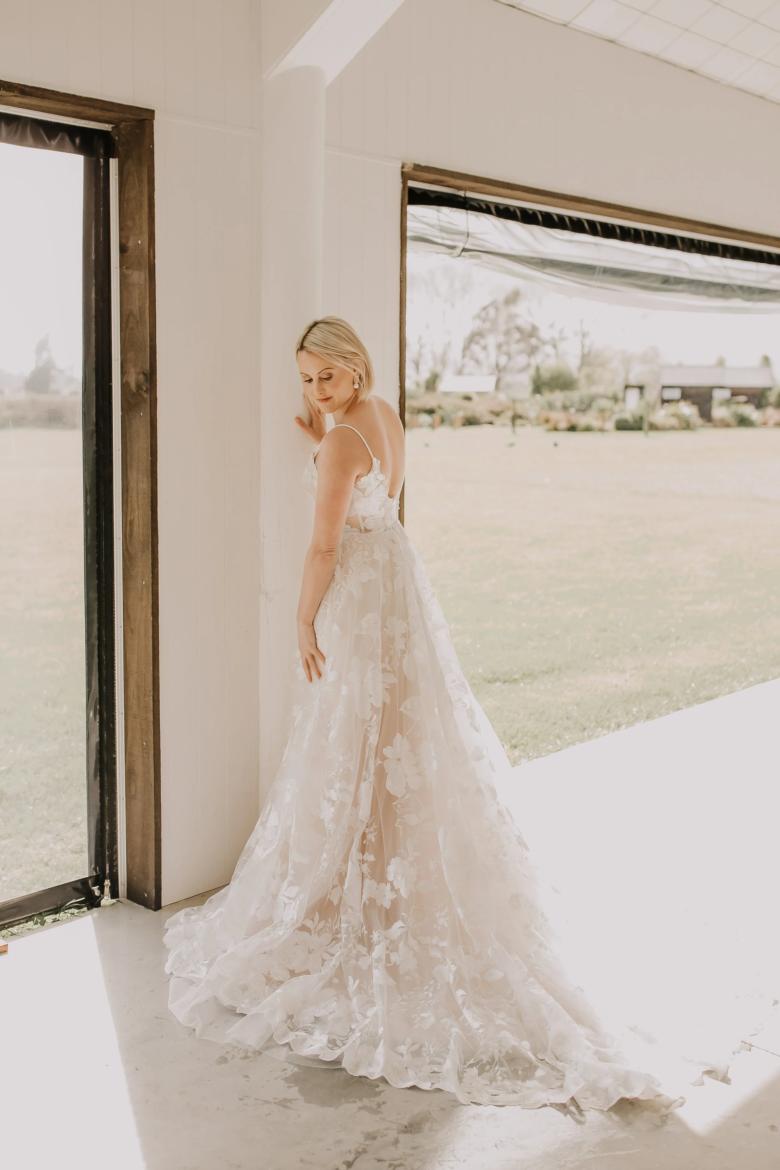 A woman in an elegant white wedding dress stands inside a bright room, gently touching a white wall near an open doorway, with a field visible outside.