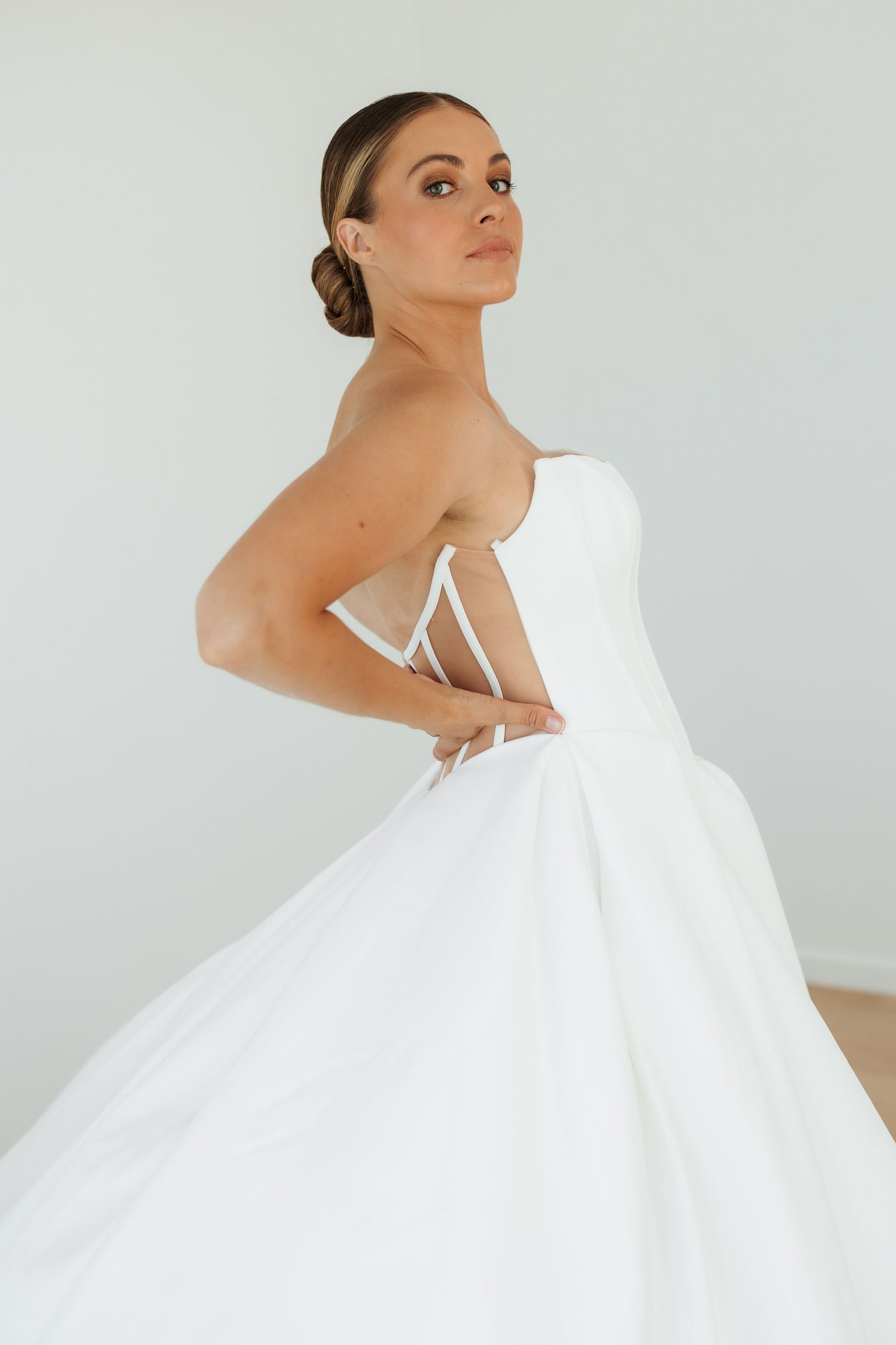 A woman in an elegant white wedding dress stands inside a bright room, gently touching a white wall near an open doorway, with a field visible outside.