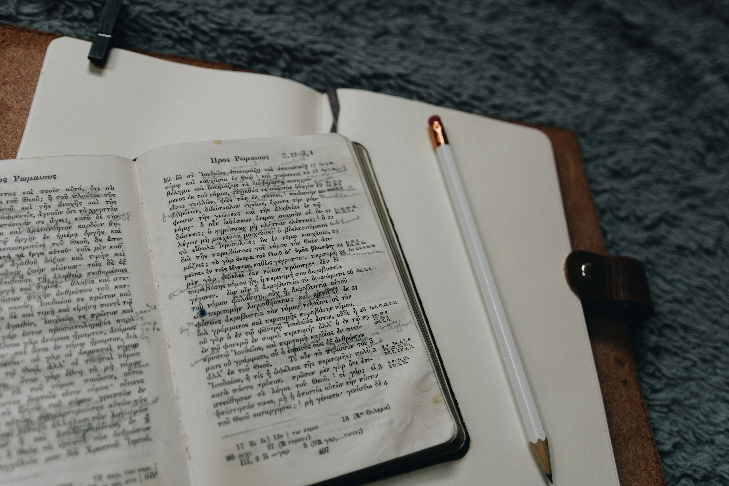 Open book with Greek text on a desk, a pencil placed next to the book, and a cloth in the background.