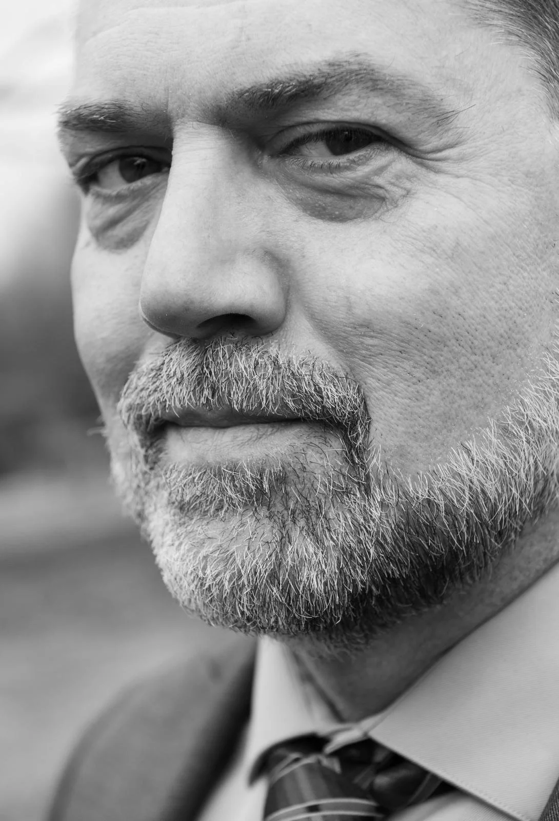Close-up black-and-white photograph of an older man with a graying beard, wearing a suit and tie, with a focus on his face and calm expression.