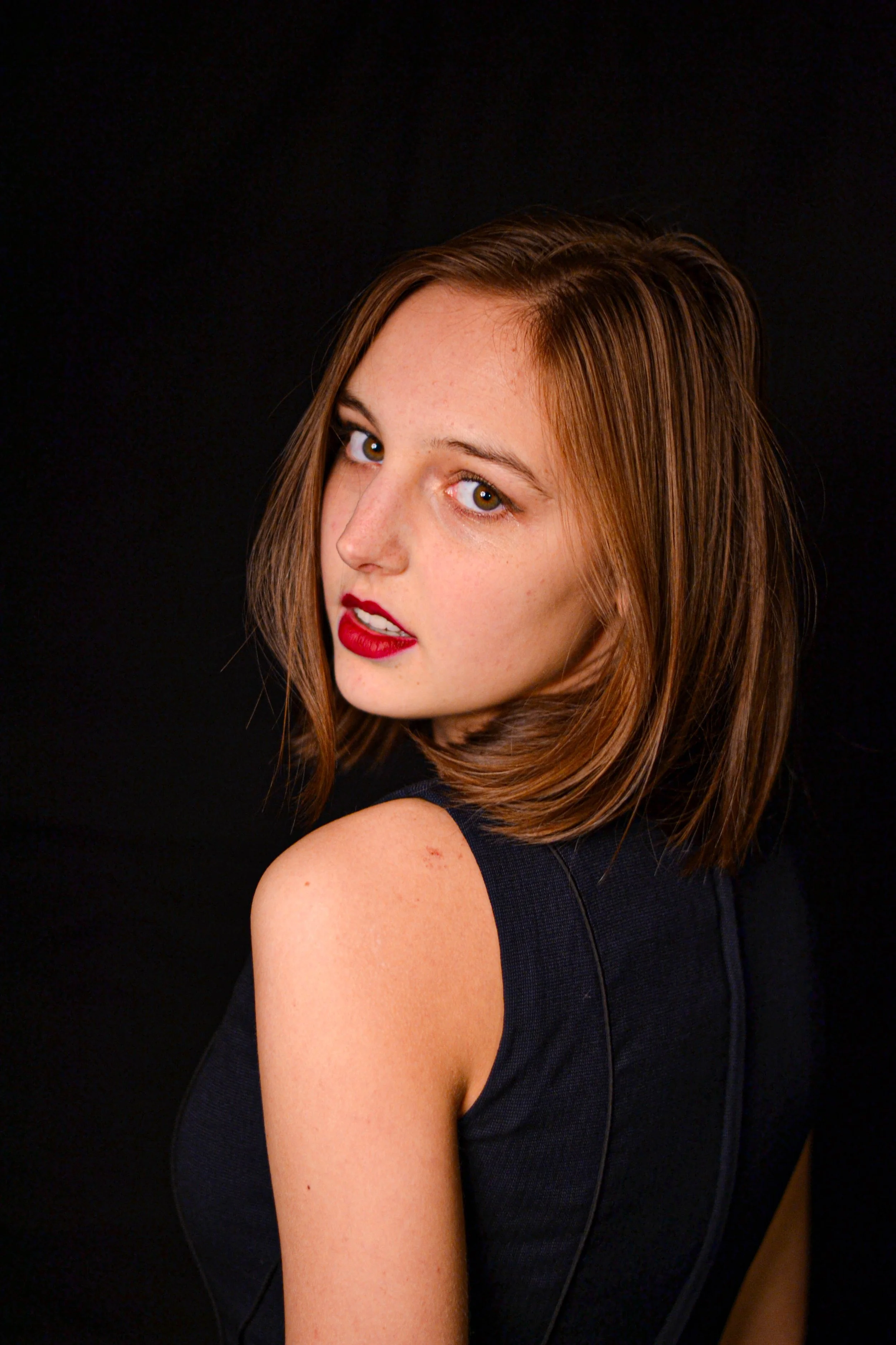 Jeune femme aux cheveux châtains, maquillée avec du rouge à lèvres rouge, portant une robe noire, contre un fond noir.