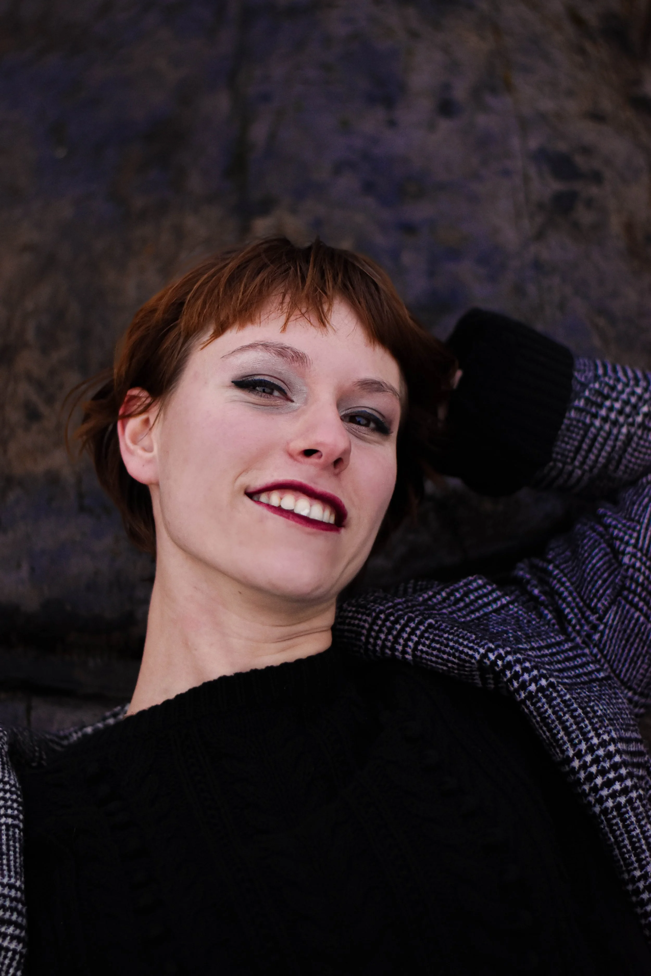 Jeune femme avec coupe de cheveux courte, rousse, souriante, portant un pull noir et un blazer à carreaux, posant à proximité d'un mur en pierre.