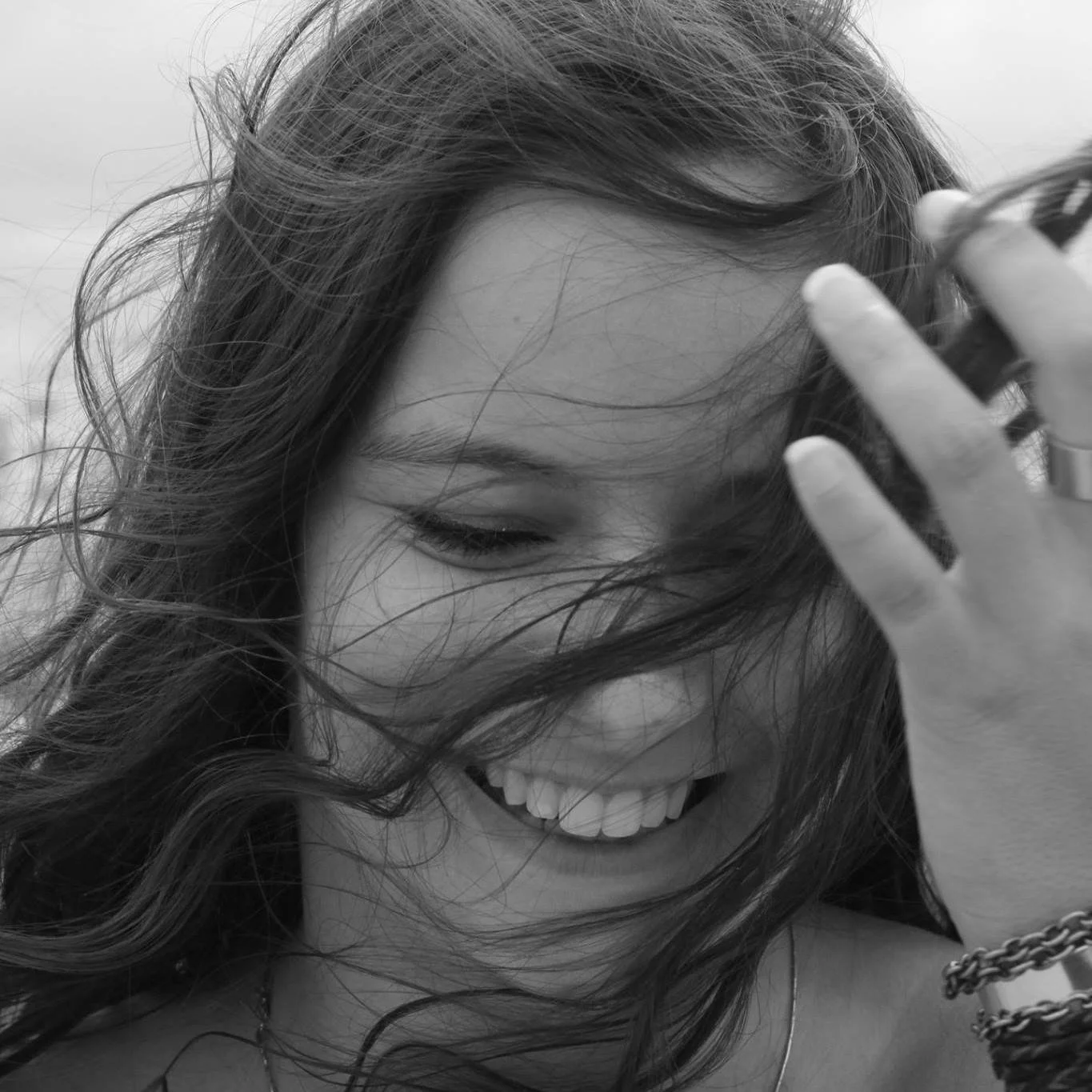 Une jeune femme souriante, les yeux fermés, replaçant ses cheveux qui couvrent partiellement son visage.