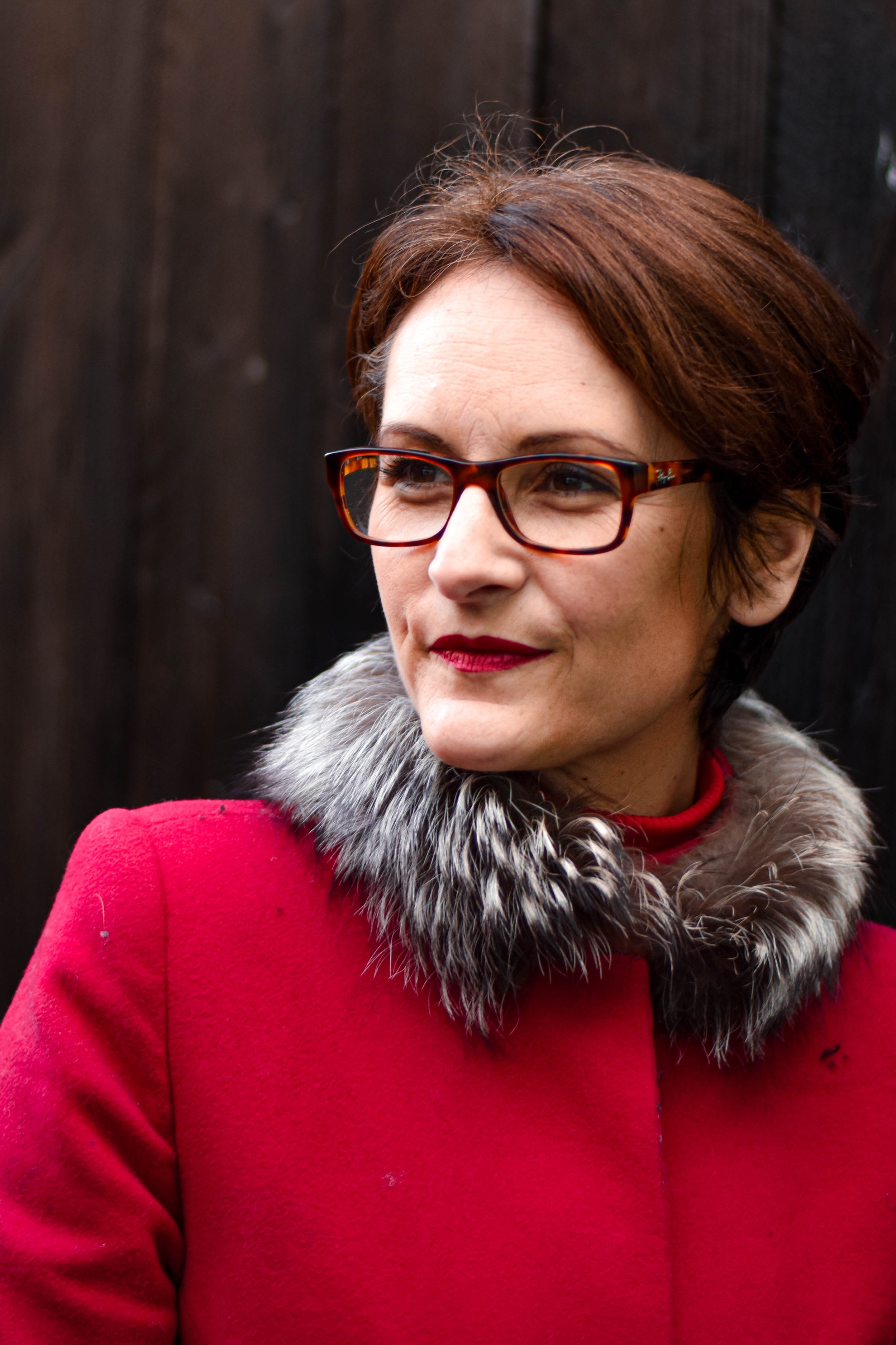 Zoom sur le visage d'une femme portant des lunettes à monture écaille, avec une coupe de cheveux courte et brune, vêtue d'un manteau rouge avec un col en fourrure gris, devant un fond sombre.