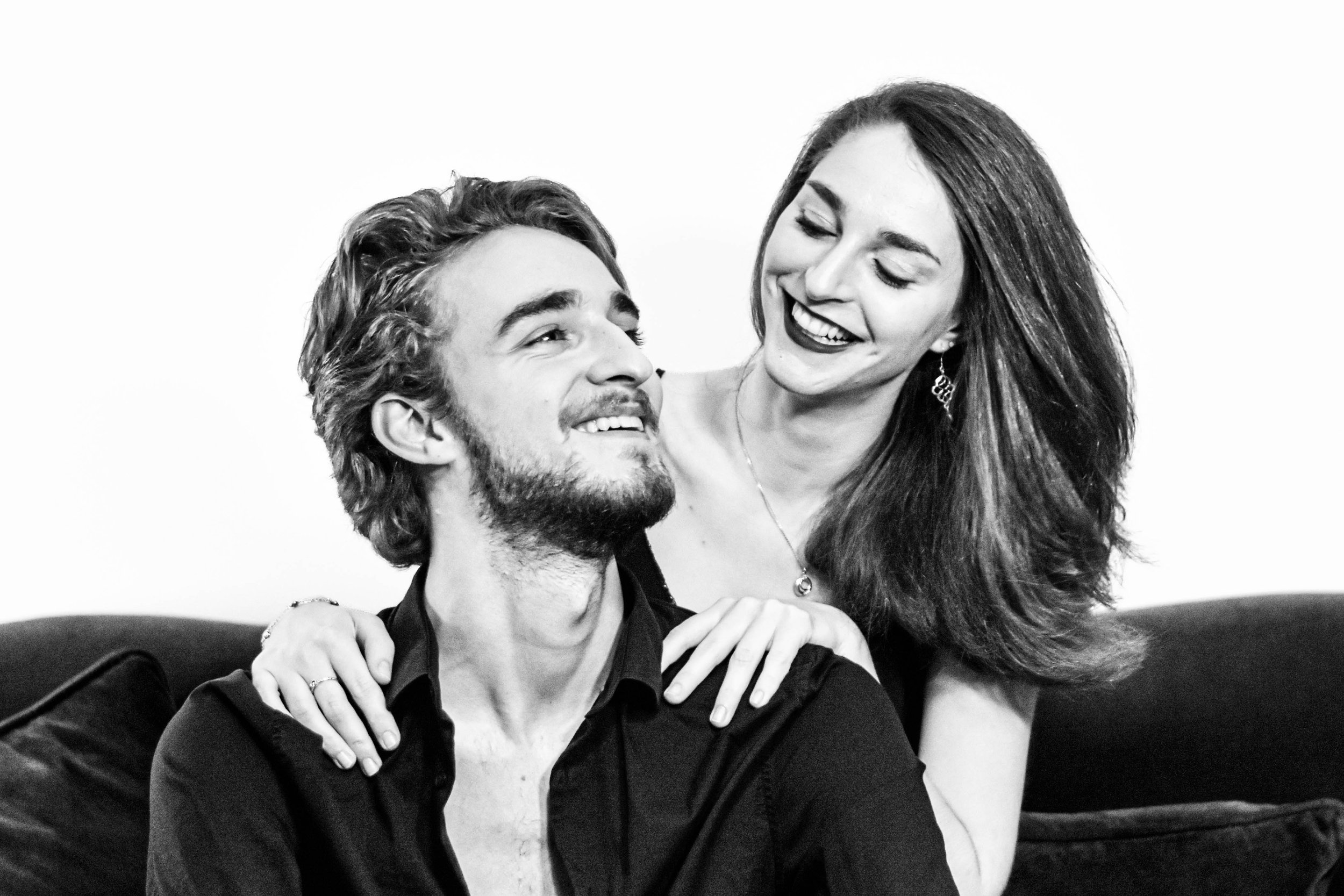 Une femme et un homme souriants, proches l'un de l'autre, dans une interaction affectueuse. L'homme porte une barbe et un T-shirt noir, la femme a de longs cheveux et porte des boucles d'oreilles étoile.