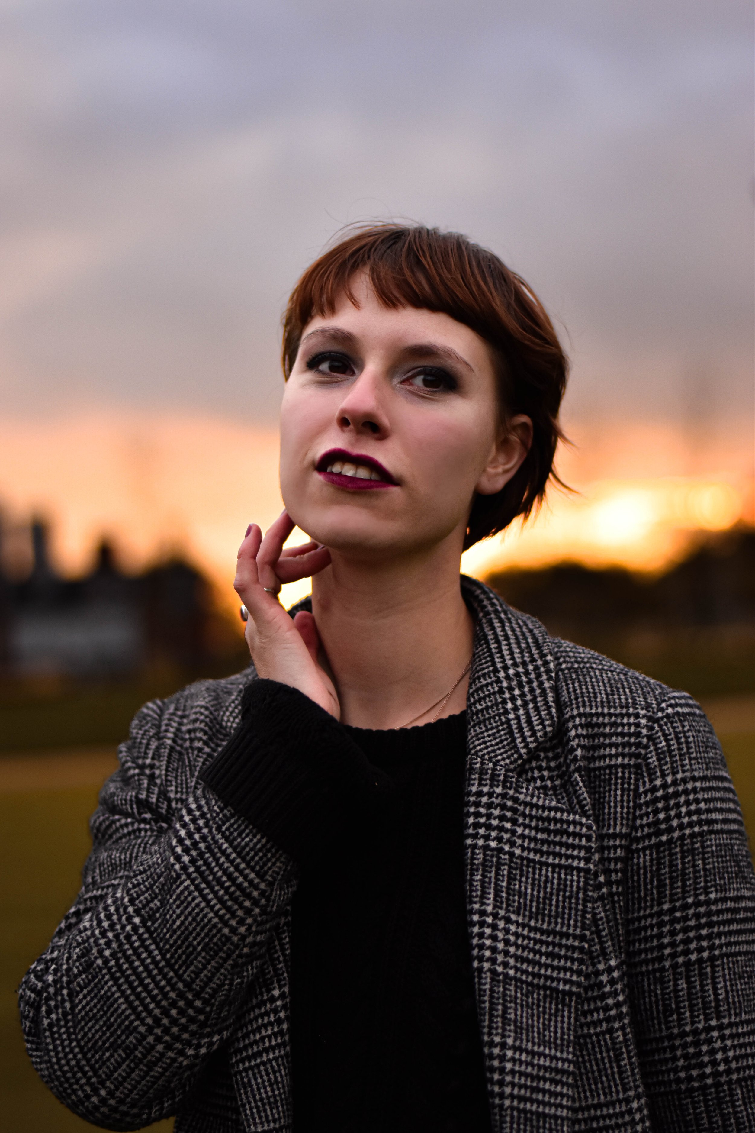 Portrait d'une jeune femme aux cheveux courts bruns, maquillée, portant un manteau à carreaux noirs et blancs, avec un fond de coucher de soleil sur un ciel nuageux.