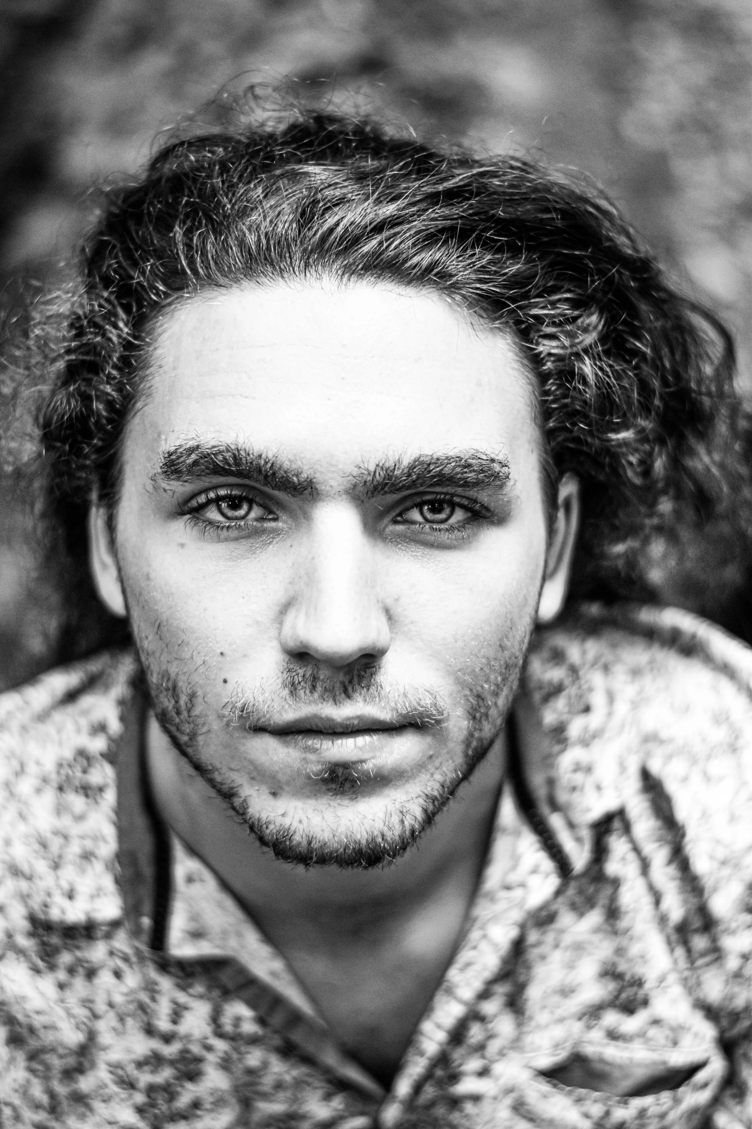 Un portrait en noir et blanc d'un jeune homme avec des cheveux bouclés, une barbe légère, regardant directement l'appareil, portant un vêtement à motif.