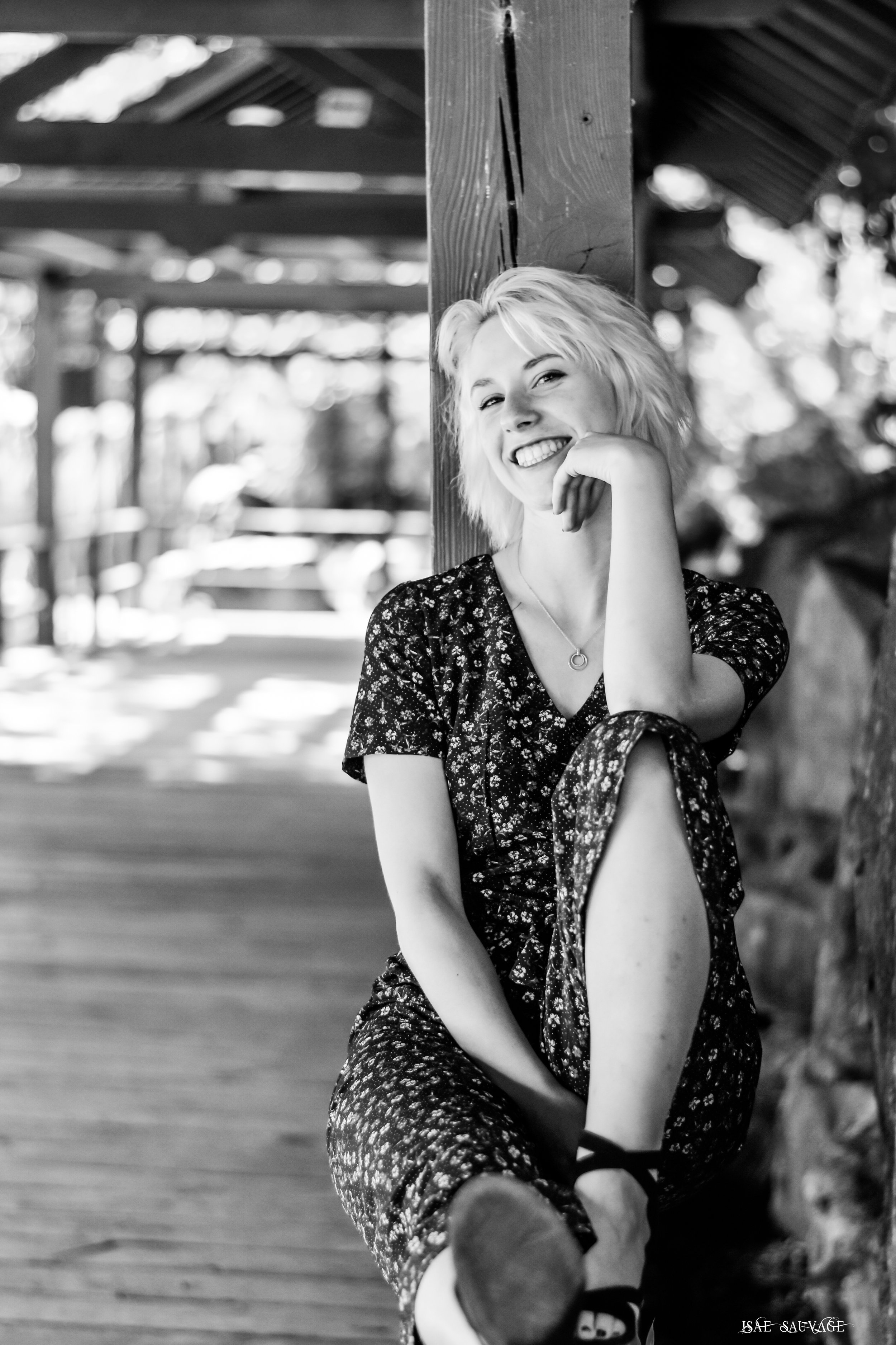 Une femme souriante, aux cheveux blonds courts, assise contre un poteau en bois dans un environnement extérieur avec une structure en bois en arrière-plan, en noir et blanc.