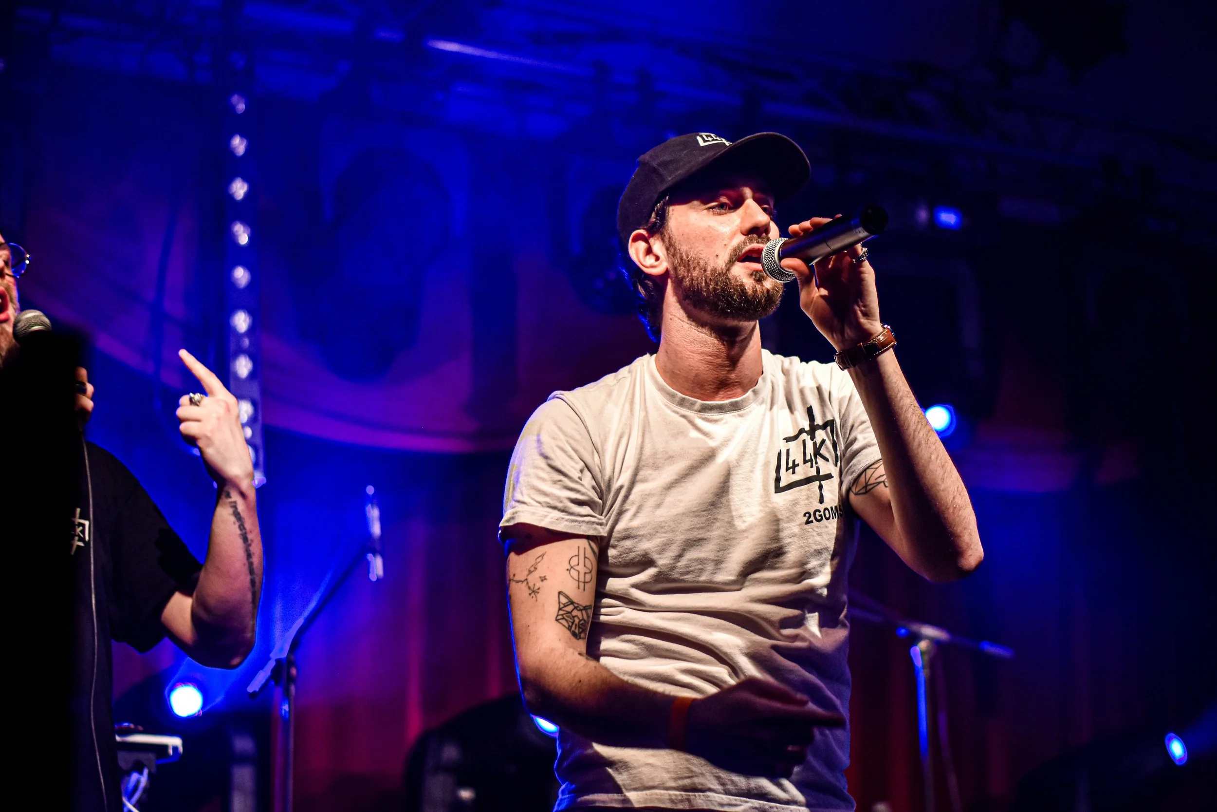 Un homme avec une barbe, portant un t-shirt blanc avec des tatouages sur les bras, tient un microphone lors d'une performance sur scène sous un éclairage violet et bleu.