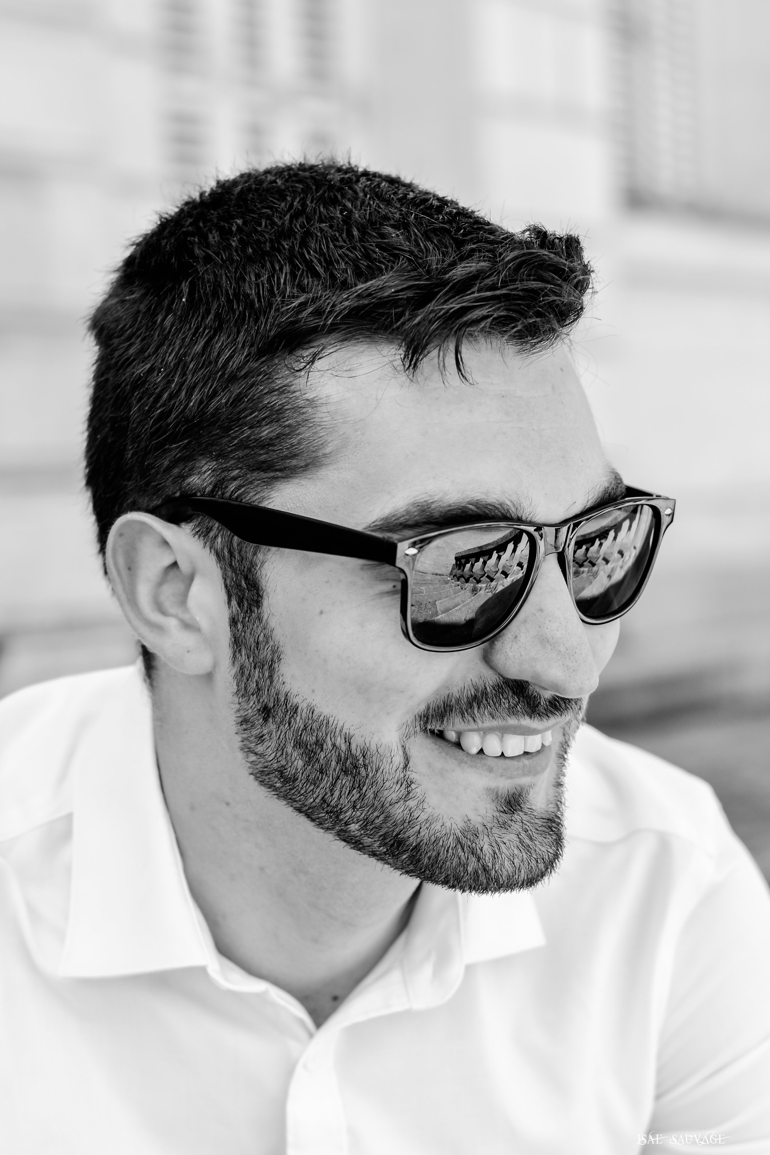 Homme souriant avec barbe, portant des lunettes de soleil, habillé d'une chemise blanche, en noir et blanc.