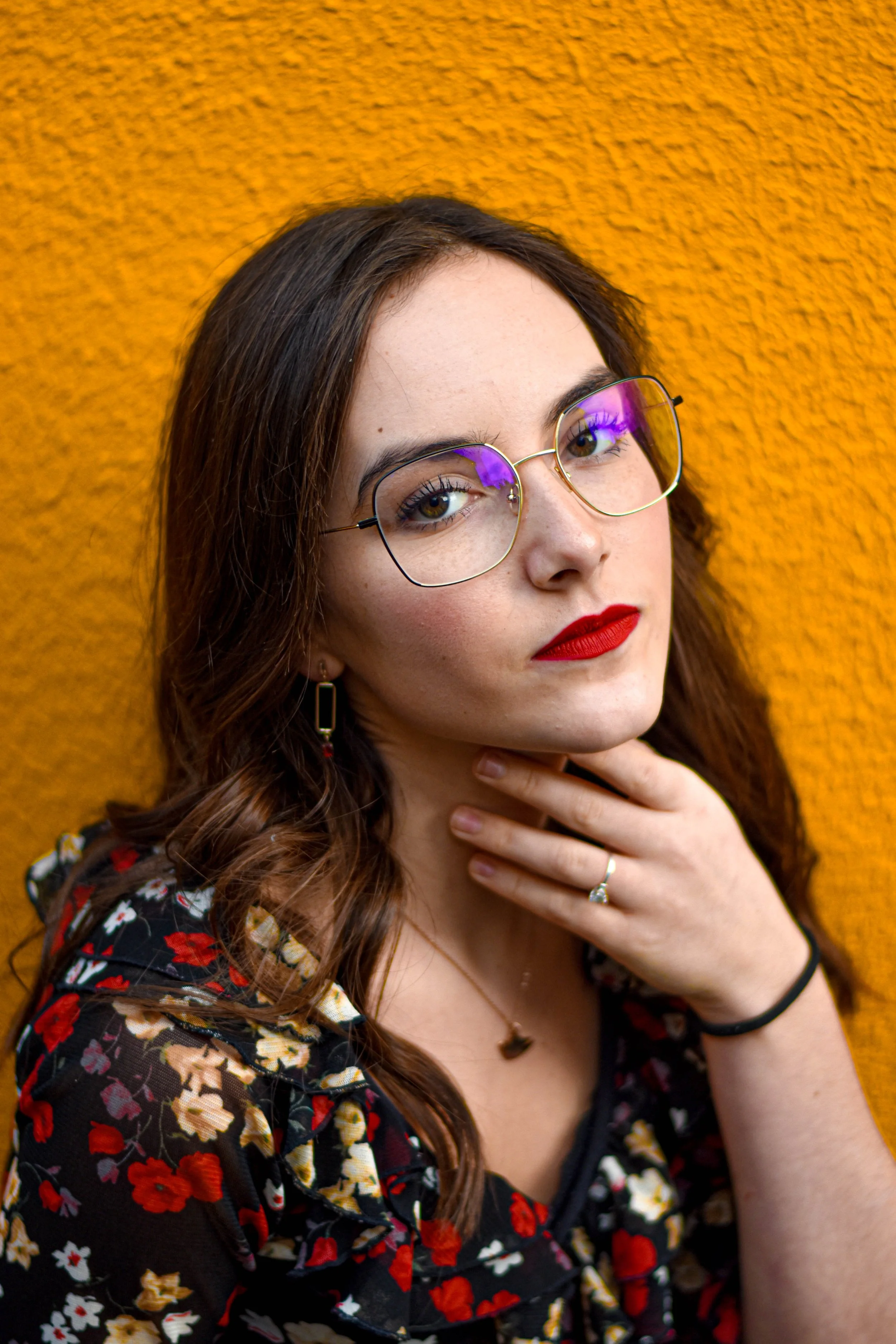 Une jeune femme avec des lunettes, portant un maquillage rouge à lèvres et un haut à fleurs, pose devant un mur orange.