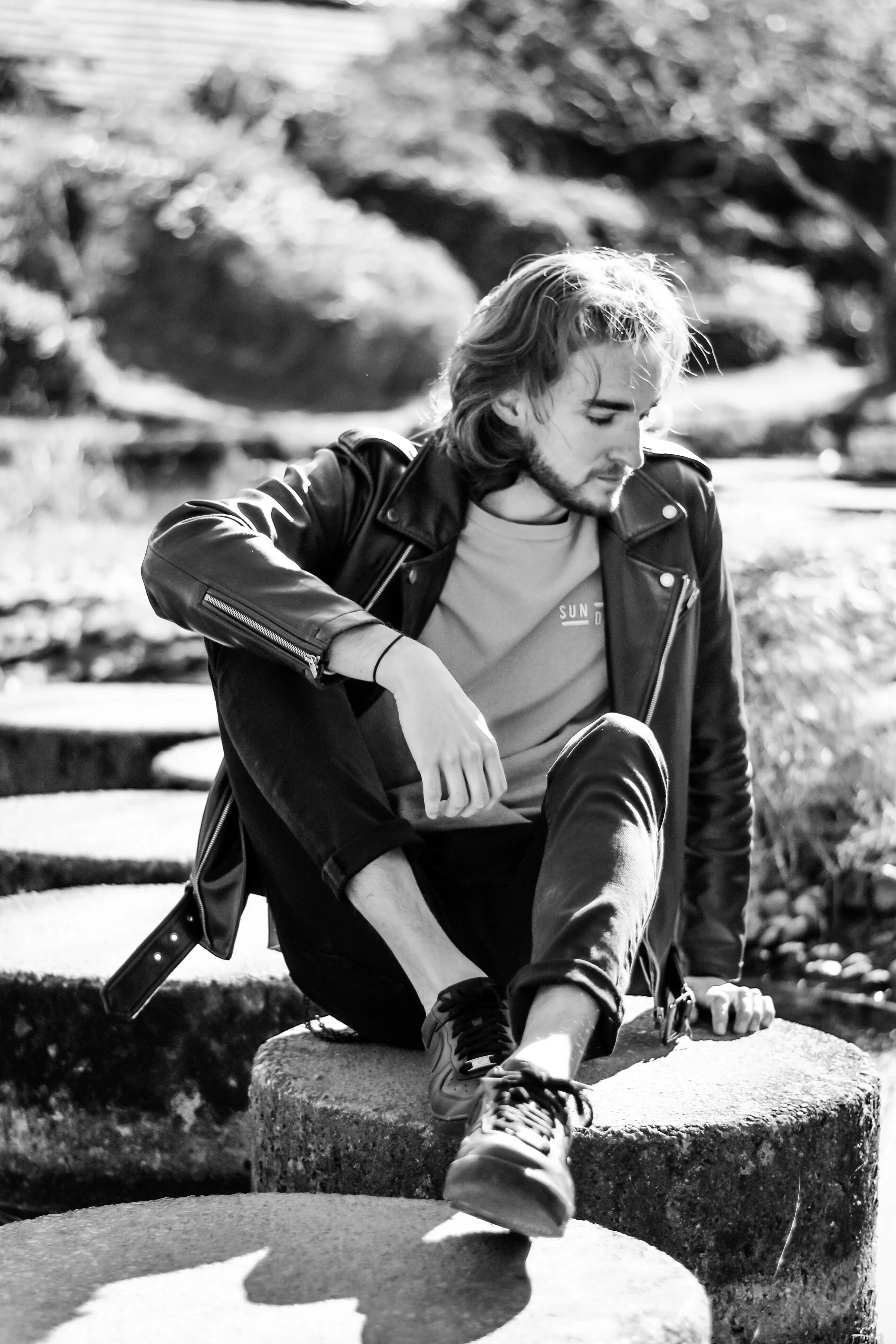 Un jeune homme avec des cheveux longs et une barbe, portant une veste en cuir, assis sur des rochers en plein air, photographié en noir et blanc.