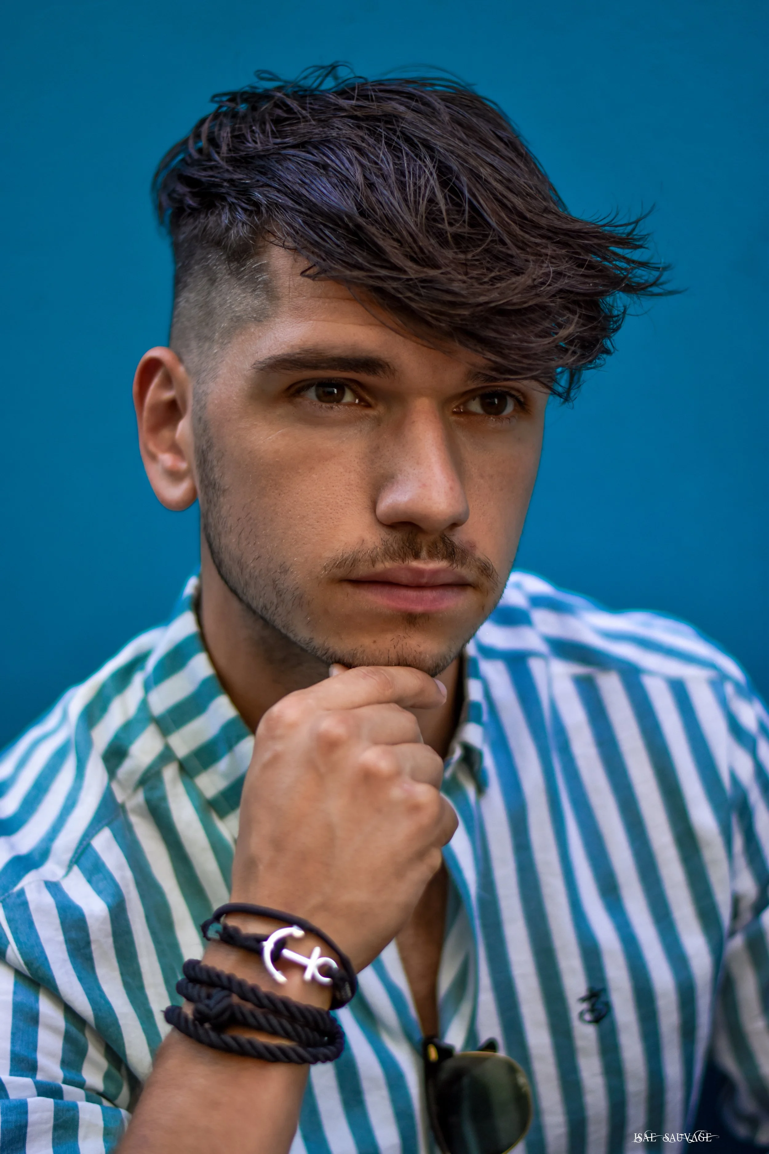 Jeune homme portant un tee-shirt à rayures bleues et blanches, posant avec la main sous le menton, porte des bracelets en corde et un bracelet avec un symbole de croix, lunettes noires accrochées à sa chemise, fond bleu.