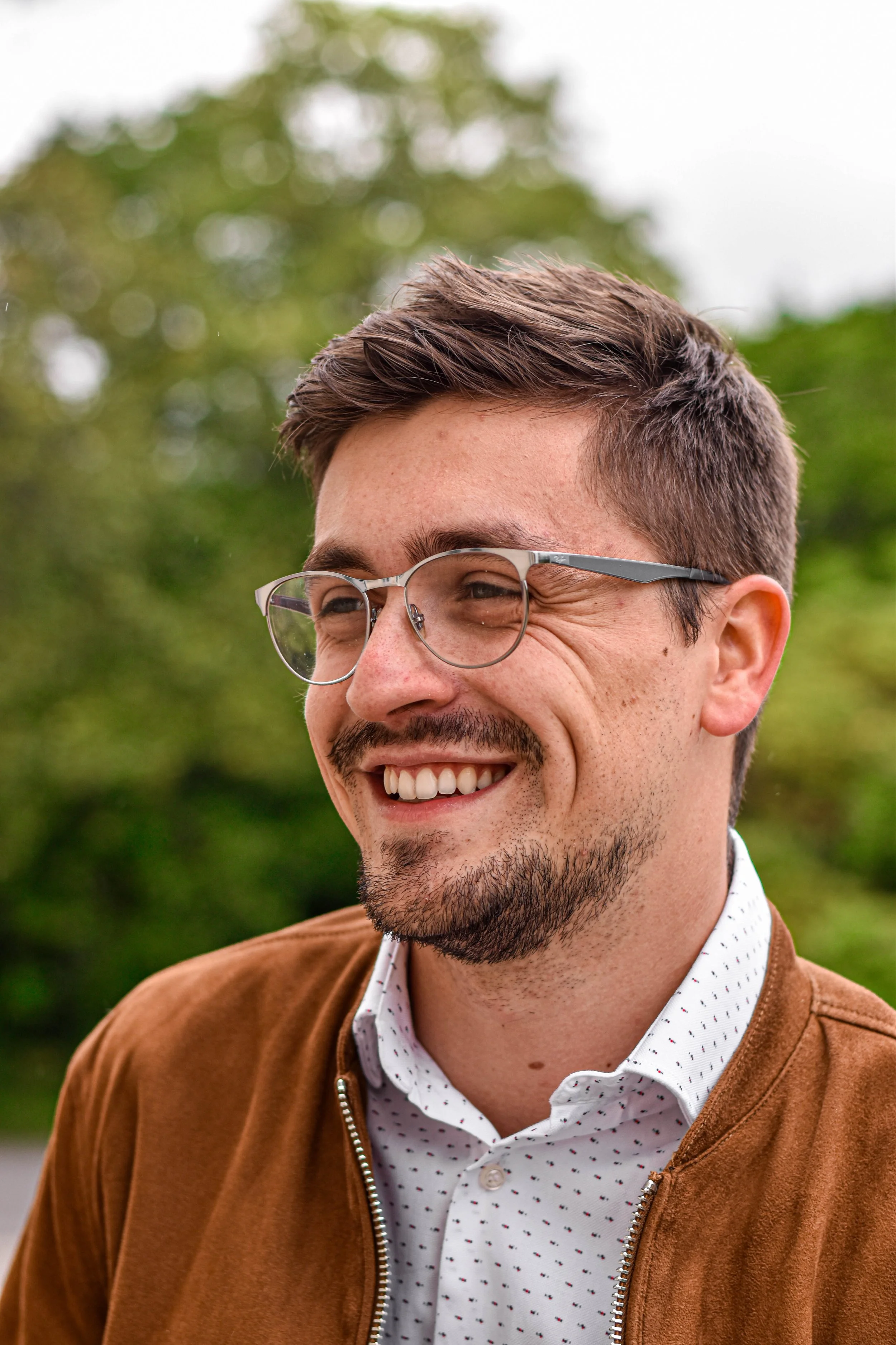 Un homme souriant portant des lunettes, une chemise blanche à pois et une veste marron, avec un fond de nature verdoyante.