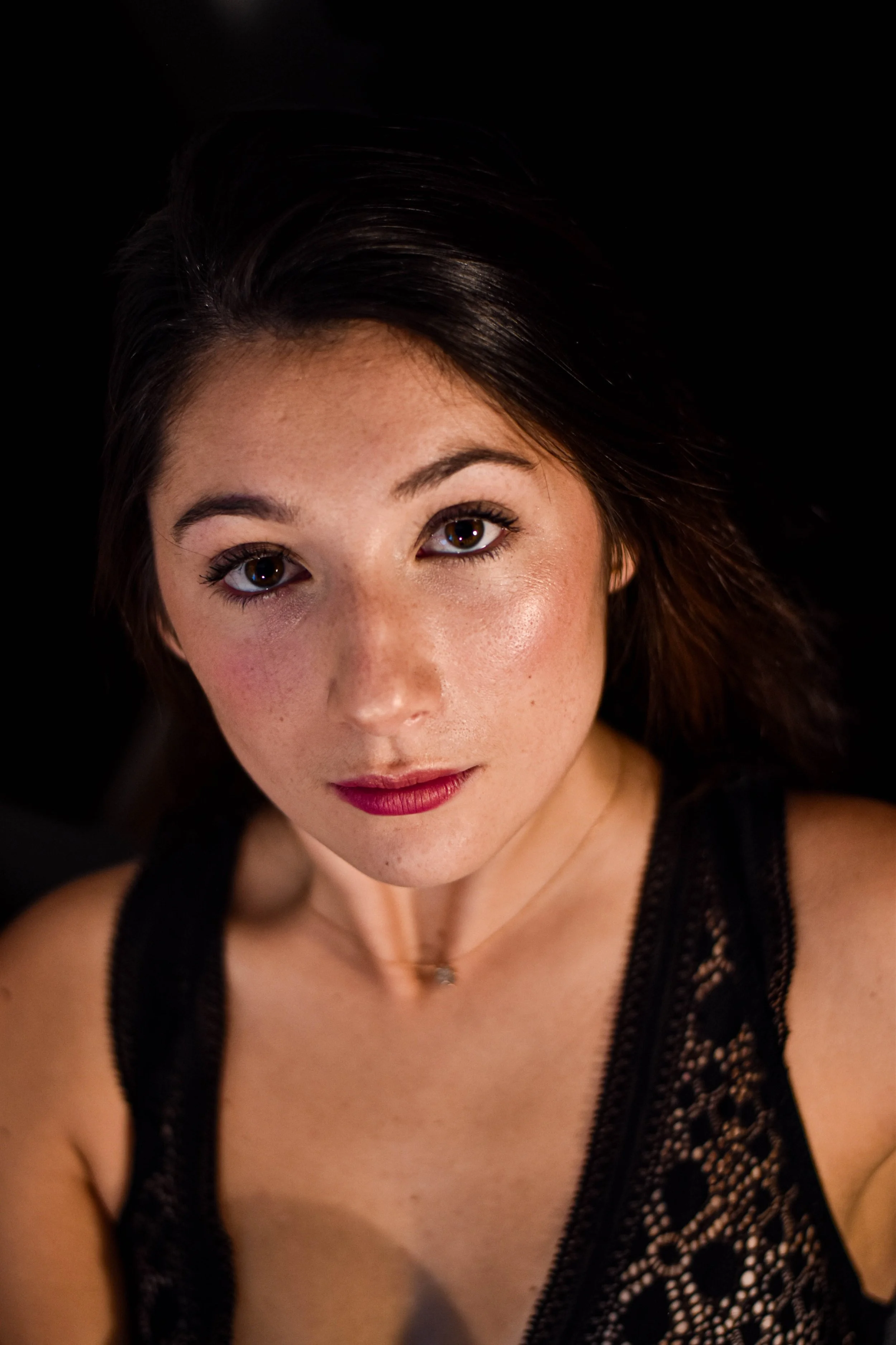 Portrait d'une jeune femme avec des cheveux bruns, portant un chemisier noir en dentelle, regardant directement l'appareil photo, fond noir.