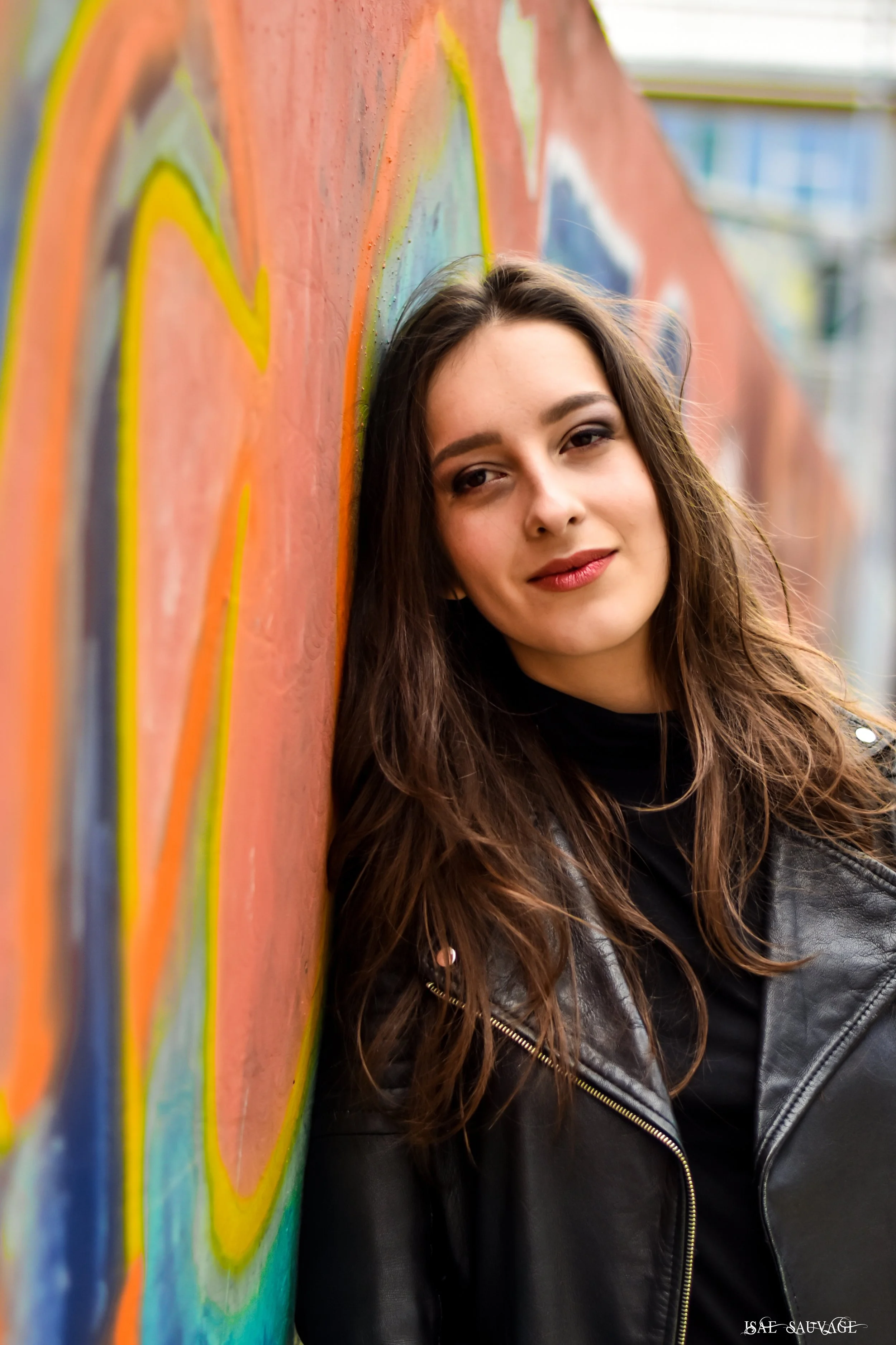 Une femme à cheveux longs et bruns avec un maquillage discret, portant une veste en cuir noir, pose contre un mur graffité coloré avec des cœurs. Elle sourit légèrement, regardant vers l'objectif.
