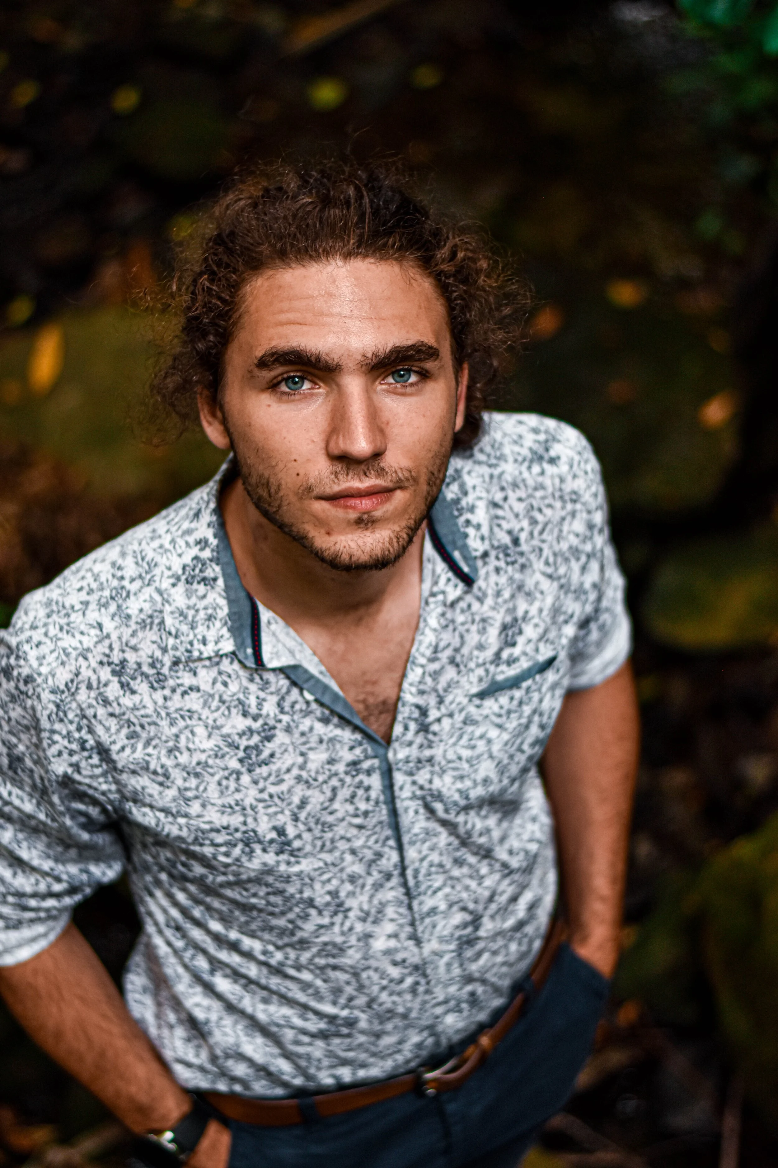 Jeune homme aux yeux bleus, avec des cheveux bouclés bruns, portant une chemise à motifs blancs et noirs, dans un environnement naturel.