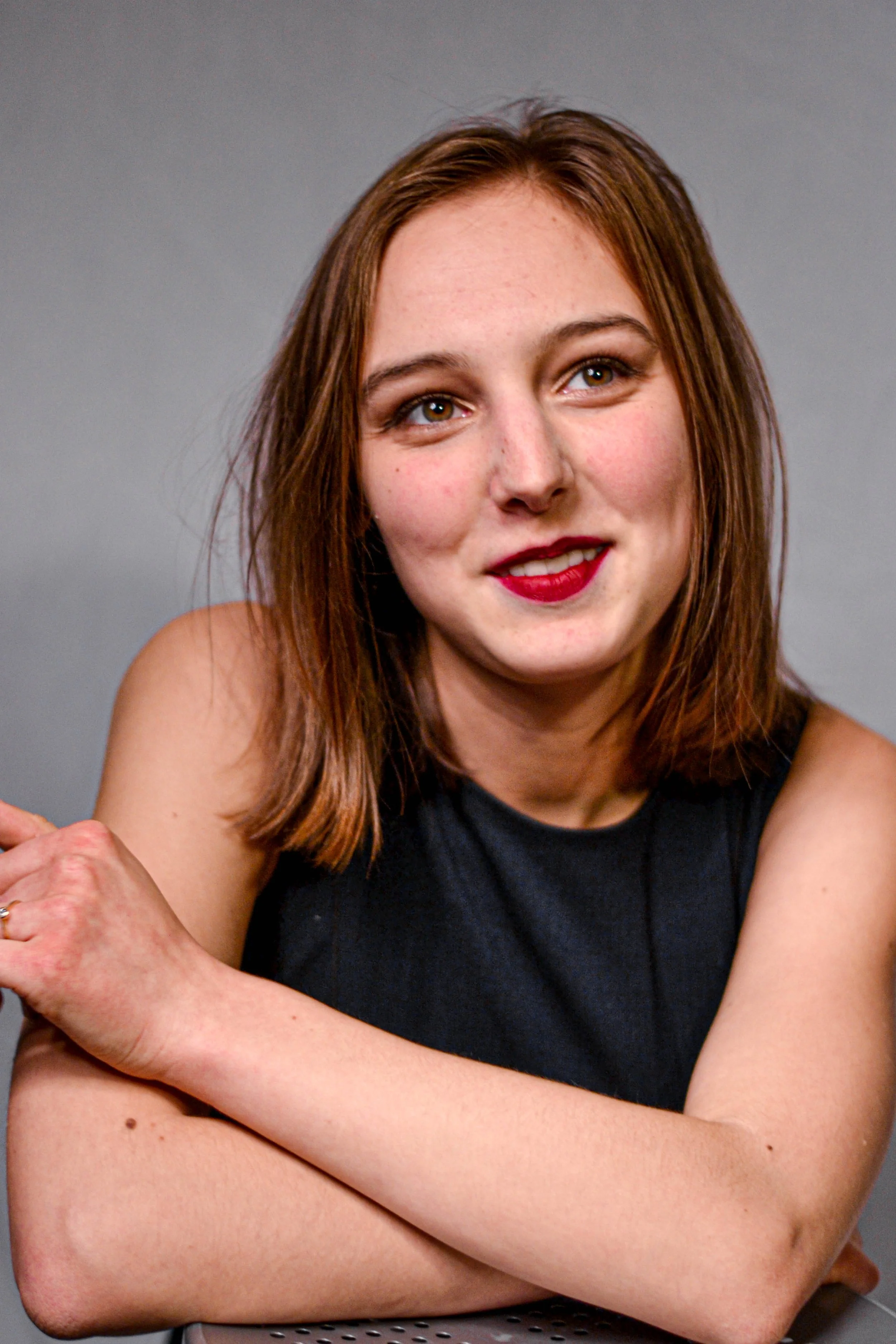 Une femme aux cheveux bruns et yeux marron, portant un haut noir, sourit légèrement et regarde vers la droite, le bras croisé contre le corps, contre un fond gris.