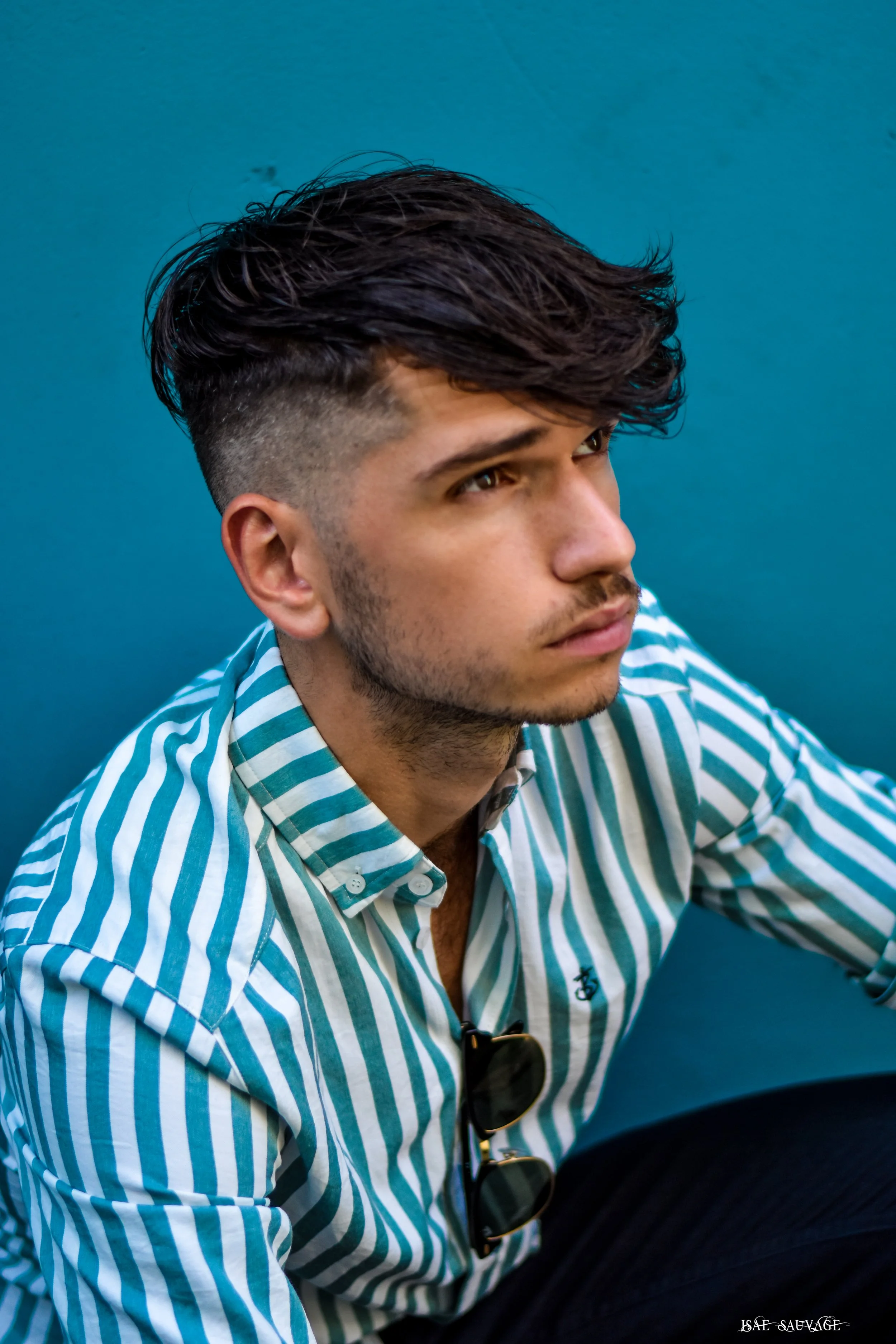 Portrait d'un jeune homme avec une coupe de cheveux courte sur les côtés et plus longue sur le dessus, portant une chemise à rayures bleu et blanc, face à droite, assis contre un mur bleu.