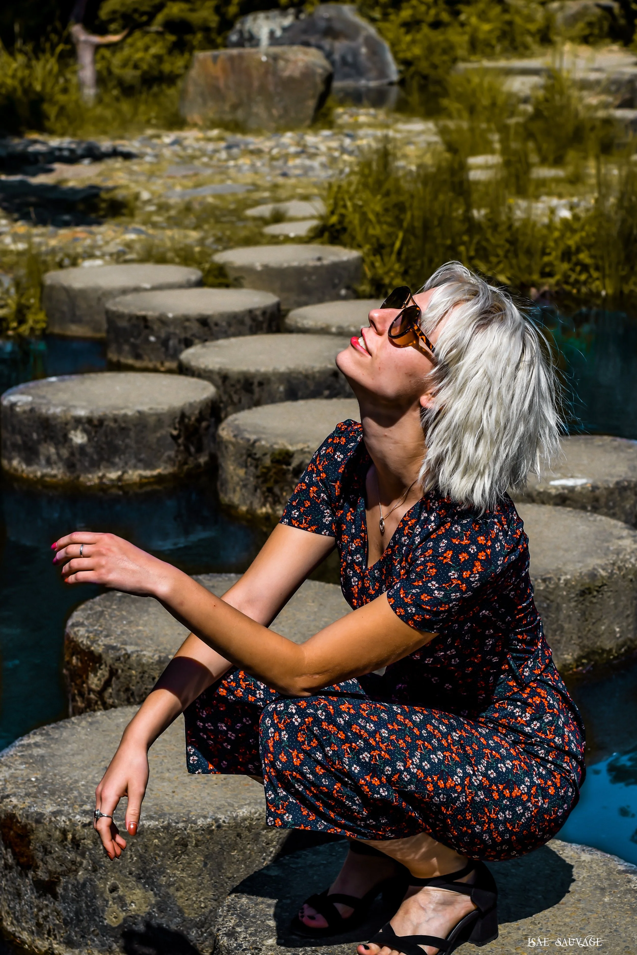 Femme blonde aux lunettes de soleil, habillée d'une robe à motif floral, assise sur des rochers près d'une rivière ou d'un étang, avec une végétation en arrière-plan, profitant du soleil.