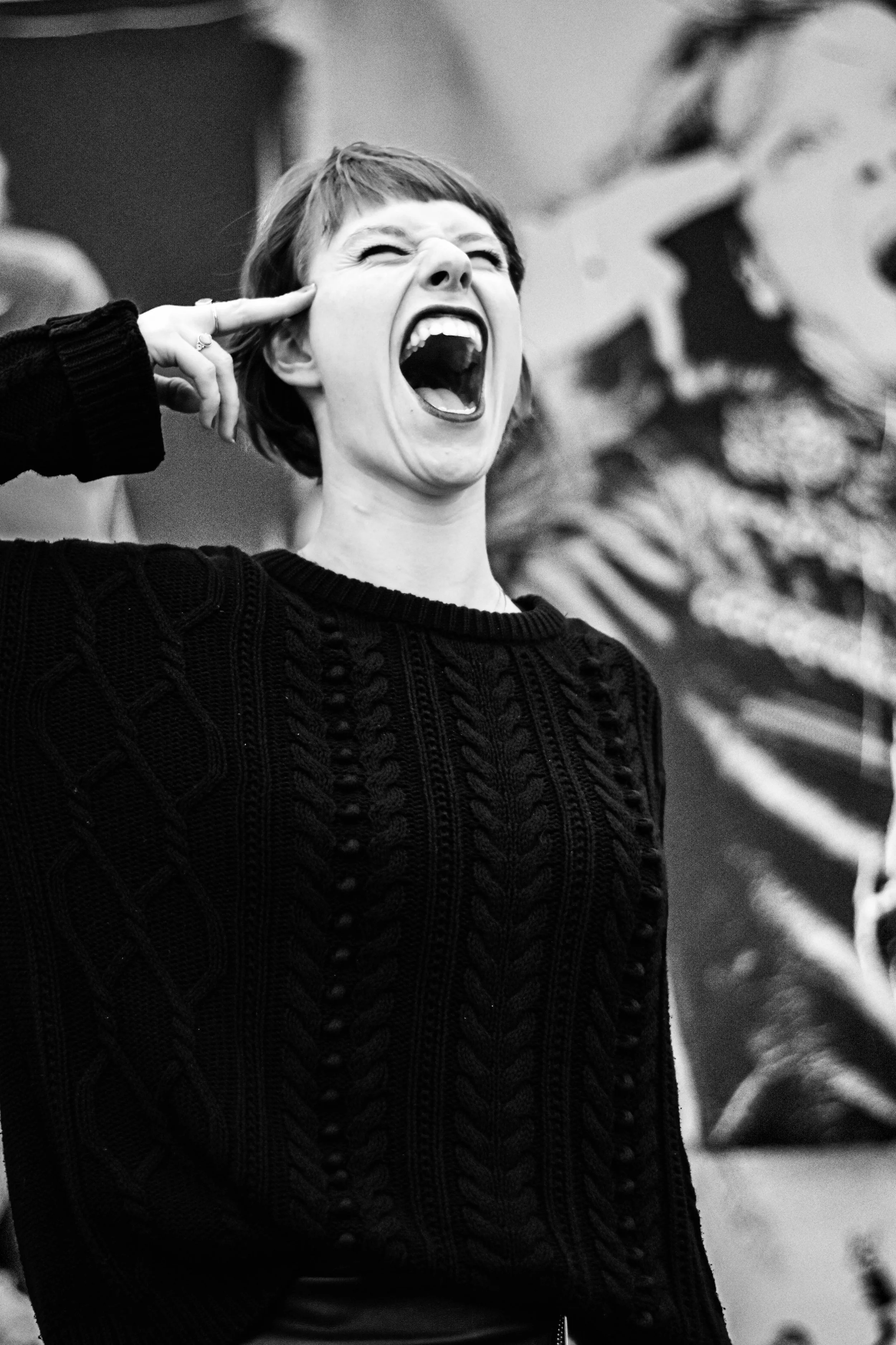 Une femme crie ou chante avec une expression intense, portant un pull en maille noire, avec un fond flou qui semble être une peinture ou une affiche artistique.