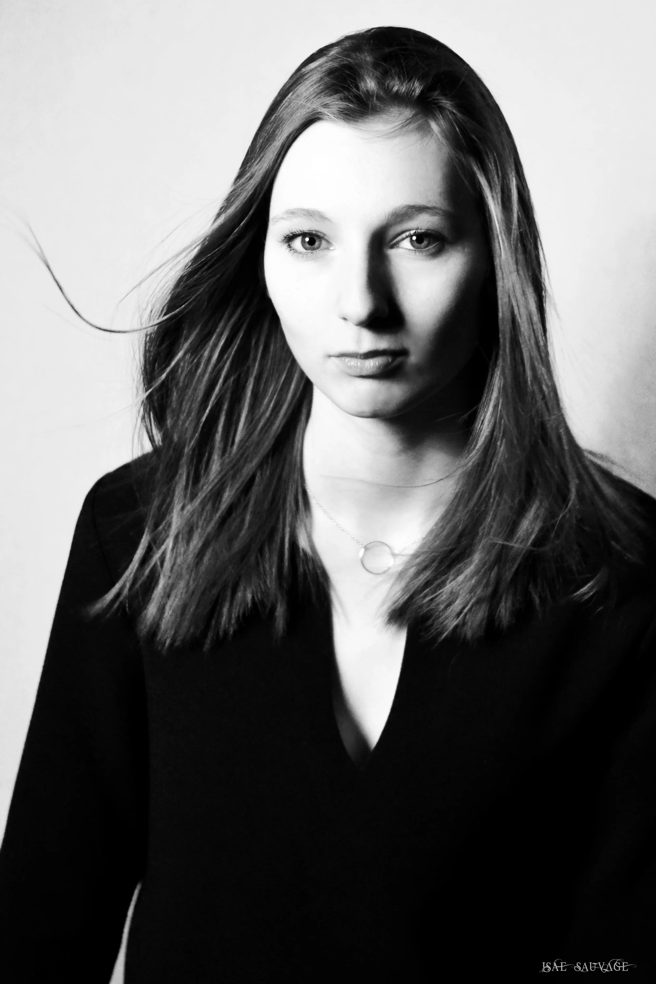 Portrait en noir et blanc d'une femme aux cheveux longs et légèrement ondulés, portant un haut noir et une fine chaîne autour du cou avec un pendentif circulaire.
