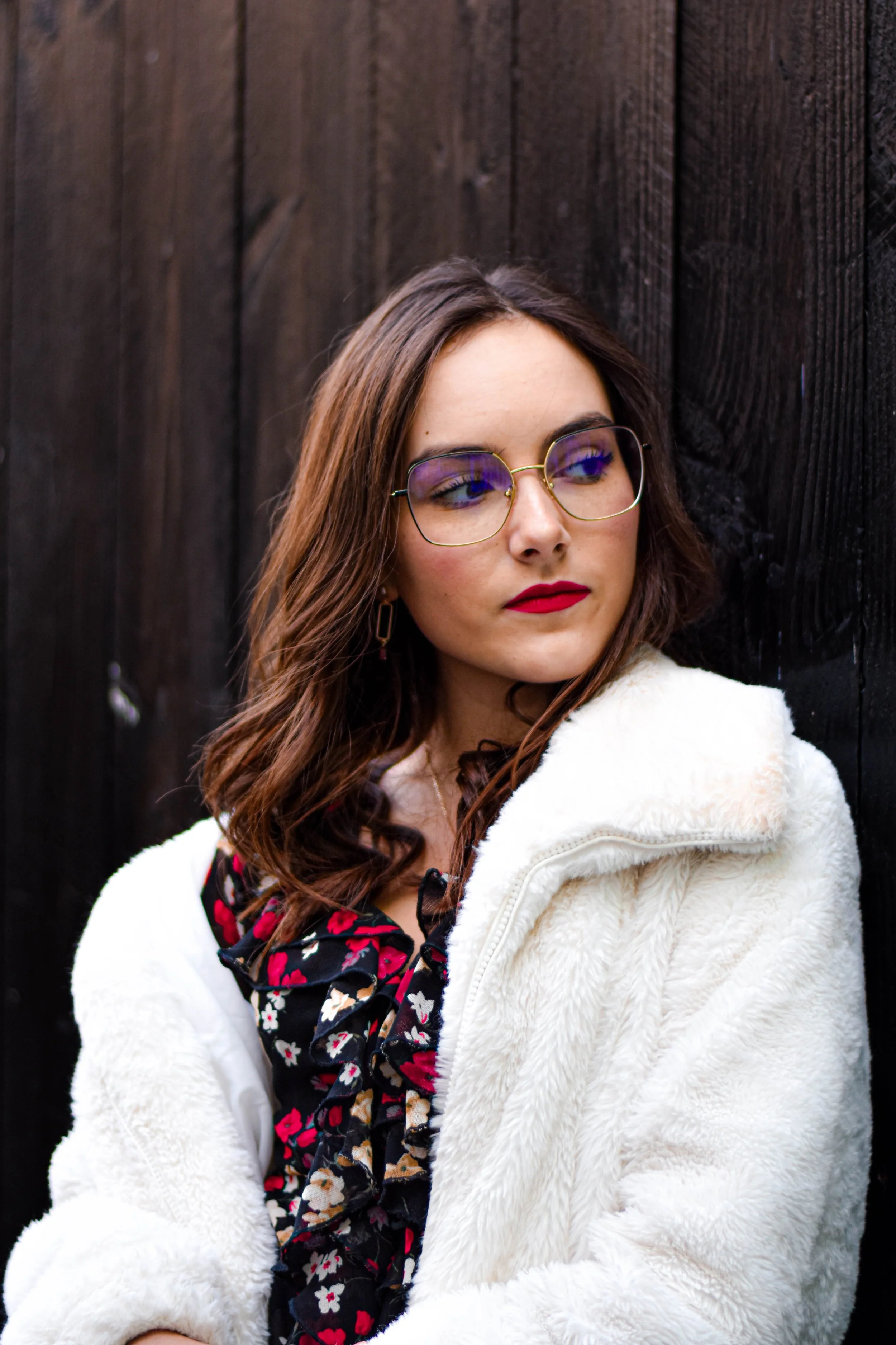 Une jeune femme avec des lunettes à monture fine porte un manteau blanc en polaire et un vêtement noir avec un motif floral rouge, blanc et beige. Elle a de longs cheveux bruns bouclés et regarde légèrement sur le côté.