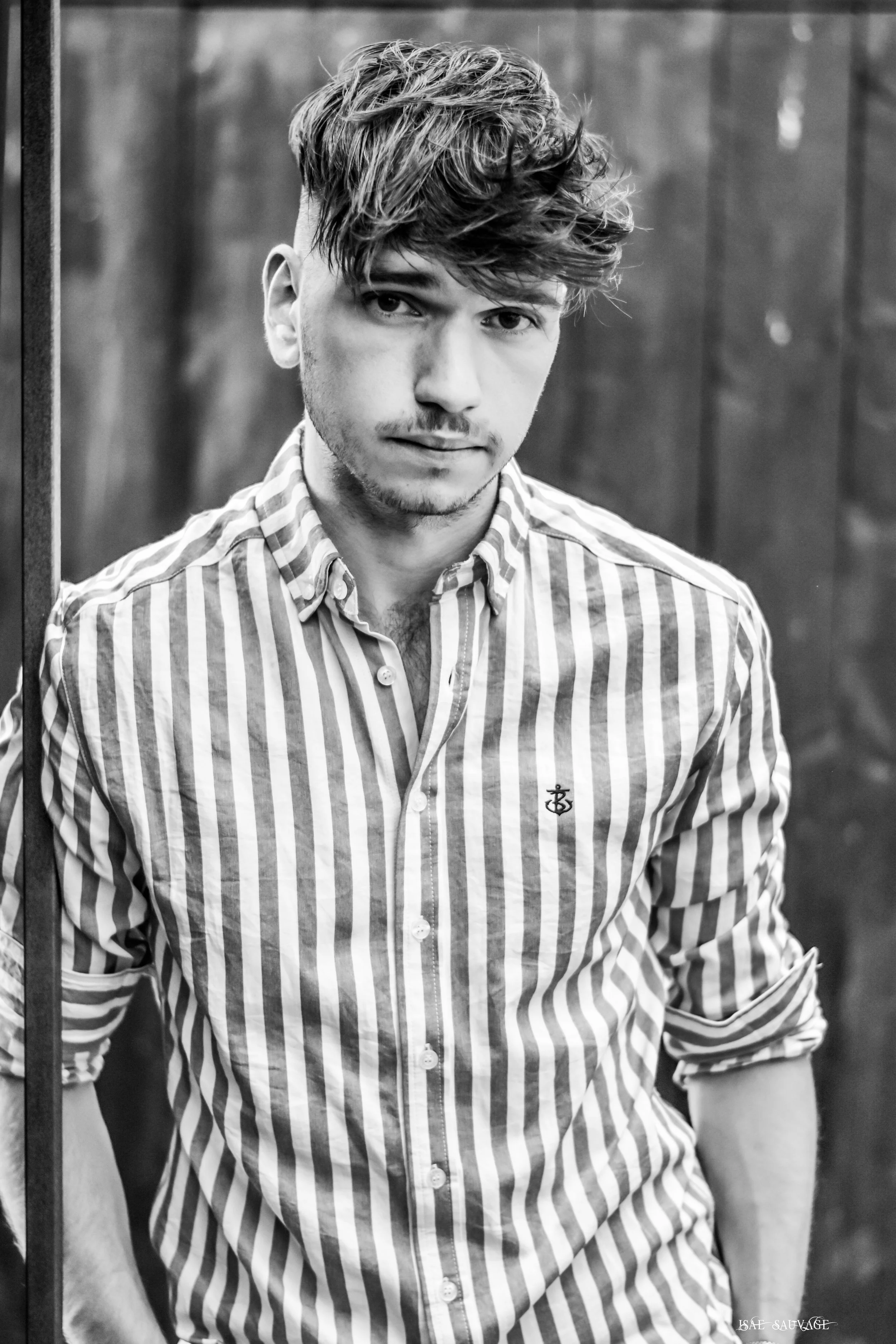 Portrait noir et blanc d'un jeune homme à cheveux bouclés portant une chemise à rayures et assis dans un lieu avec un mur en bois derrière lui.