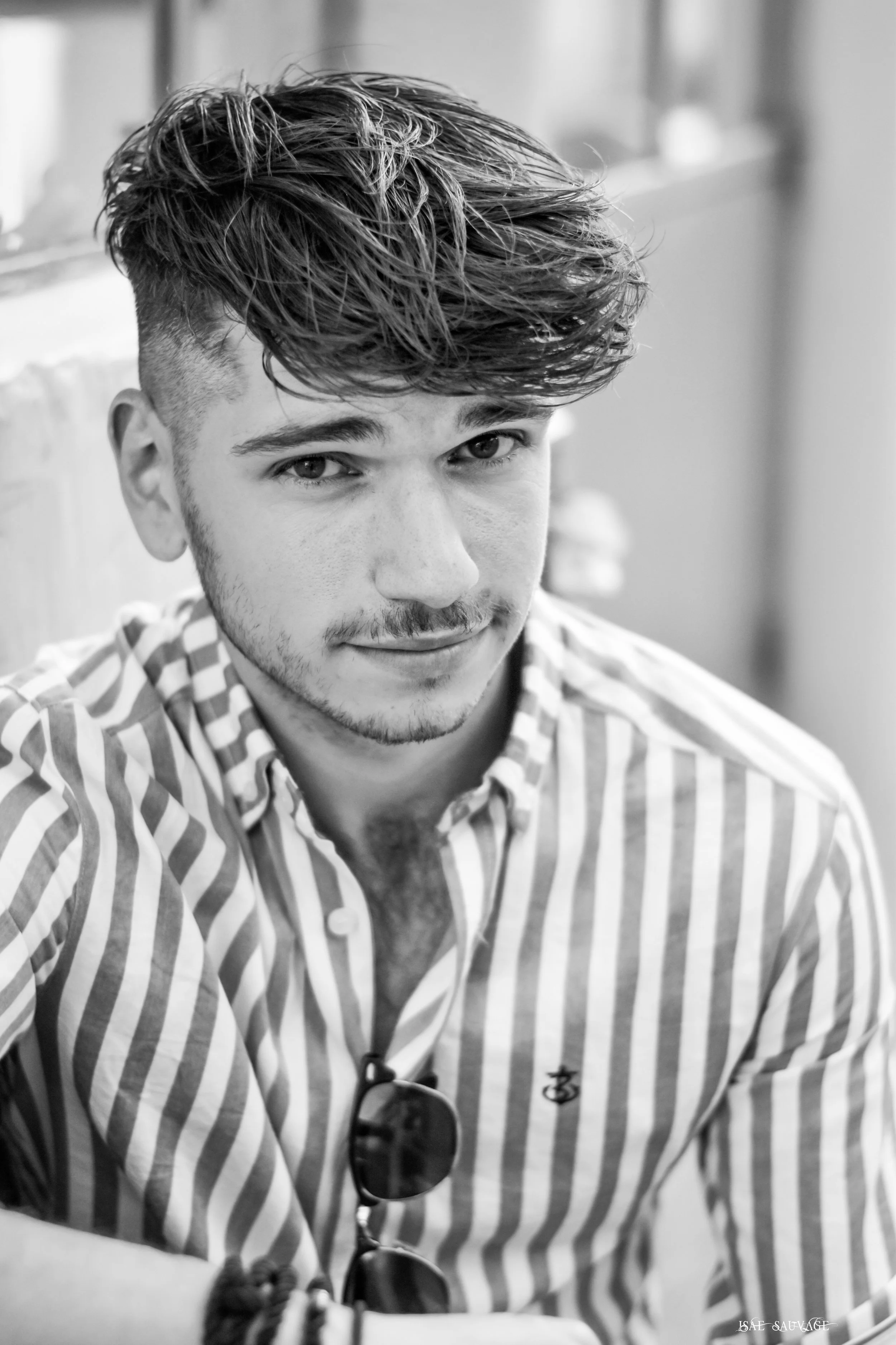 Un jeune homme avec une coiffure courte sur les côtés et une chevelure bouclée longue sur le dessus, portant des lunettes de soleil suspendues à sa chemise à rayures, regardant vers l'objectif, dans un cadre intérieur.