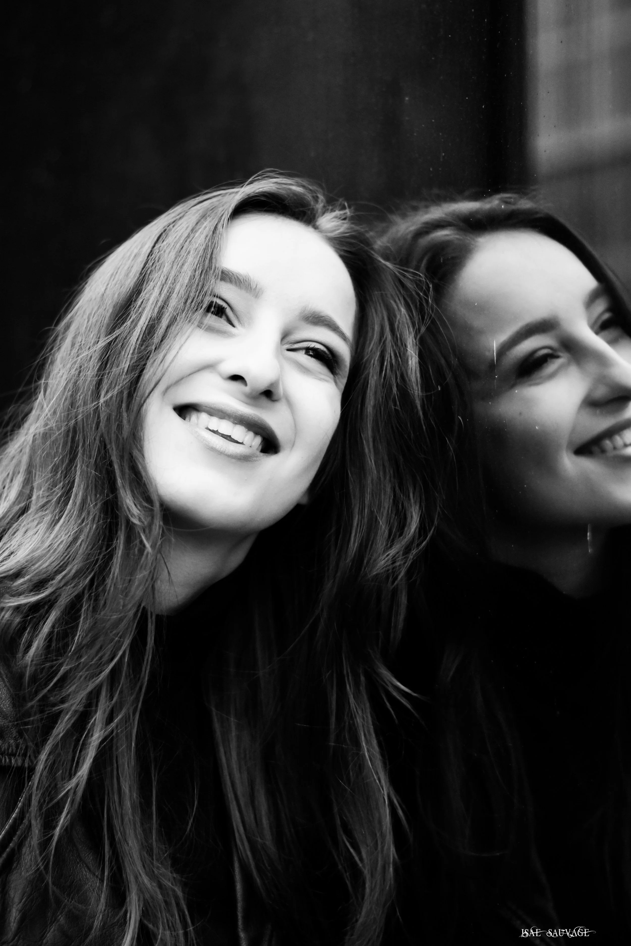 Photo en noir et blanc d'une femme souriante avec des cheveux longs, vue partielle de son visage et de sa tête, reflet dans un vitre.