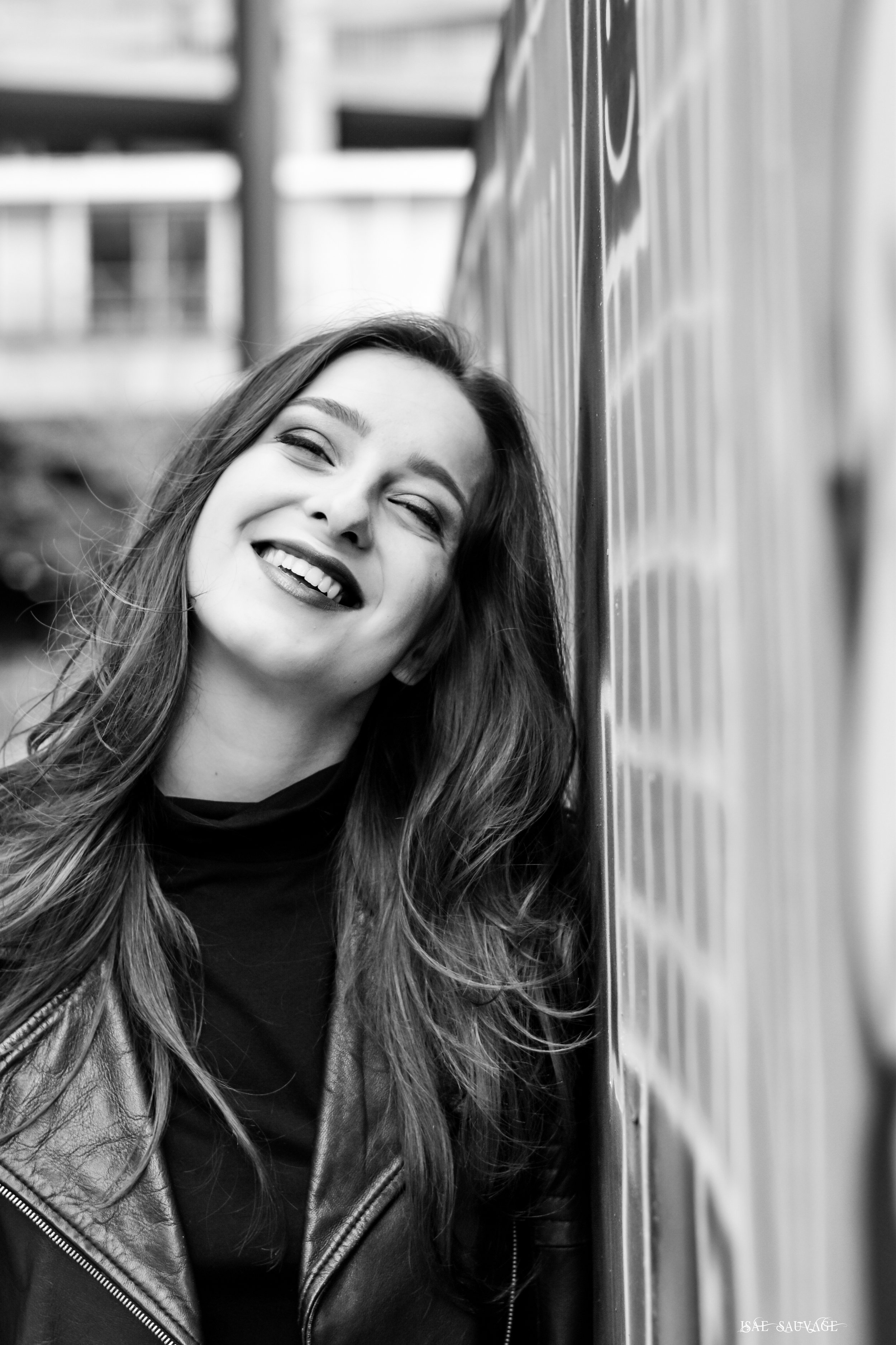 Un portrait en noir et blanc d'une femme souriante avec des cheveux longs, portant un t-shirt noir et une veste en cuir, se tenant contre une surface réfléchissante dans ce qui semble être un ascenseur ou un couloir.