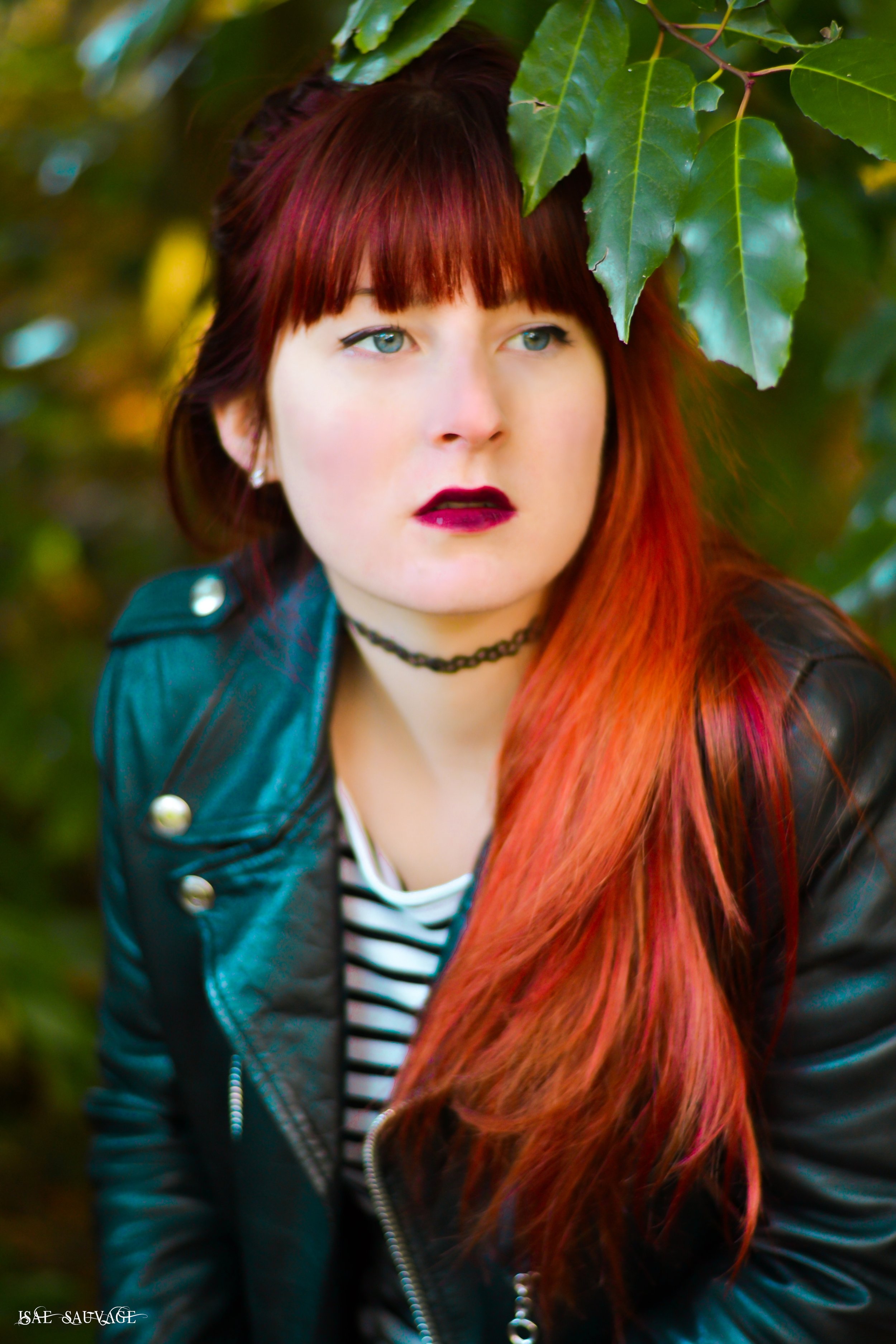 Portrait d'une femme aux cheveux longs et roux, portant une veste en cuir et un t-shirt à rayures, se tenant parmi des feuilles vertes.