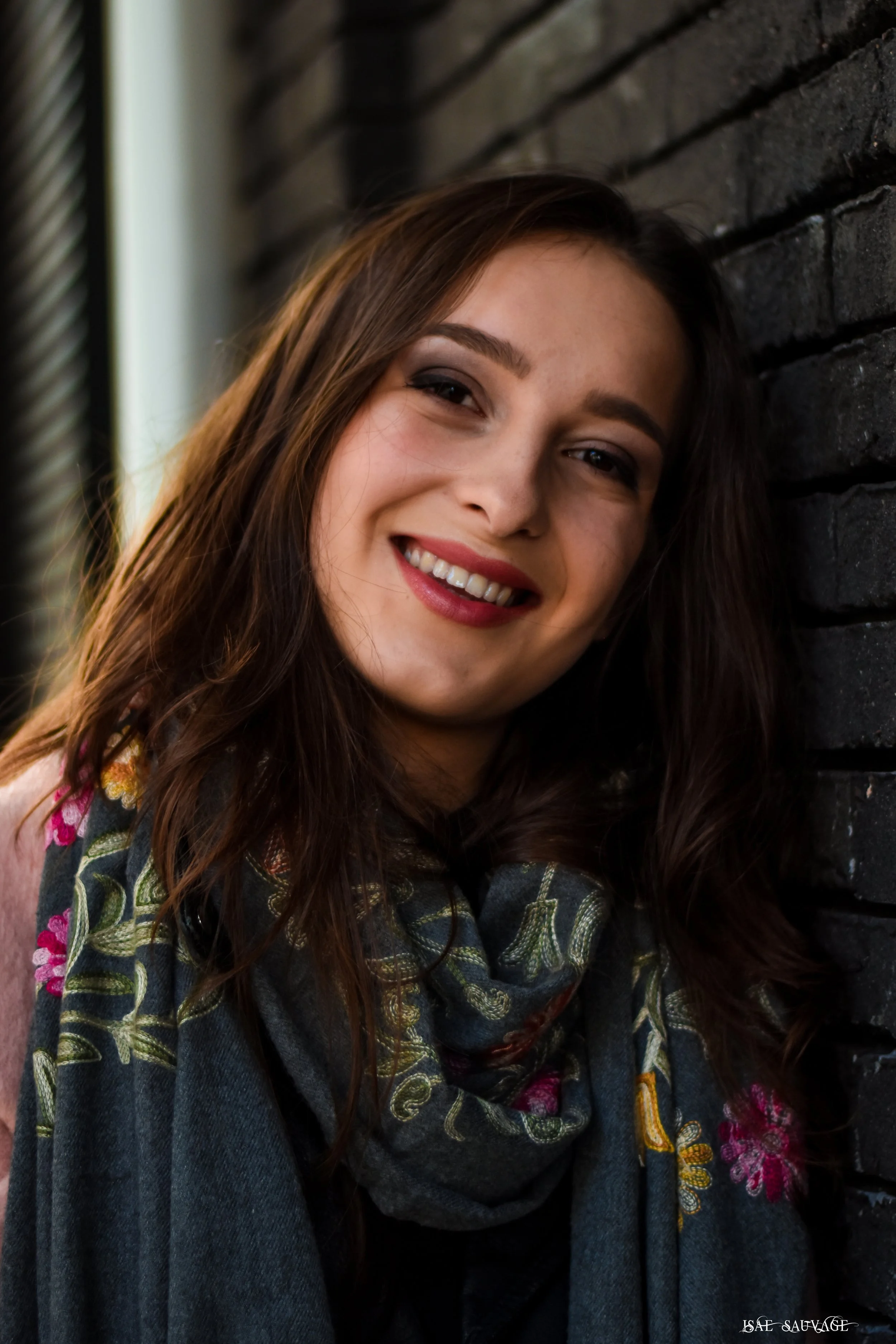 Jeune femme souriante avec cheveux longs, portant un vêtement à motifs floraux et un foulard gris, posant contre un mur en briques noires.