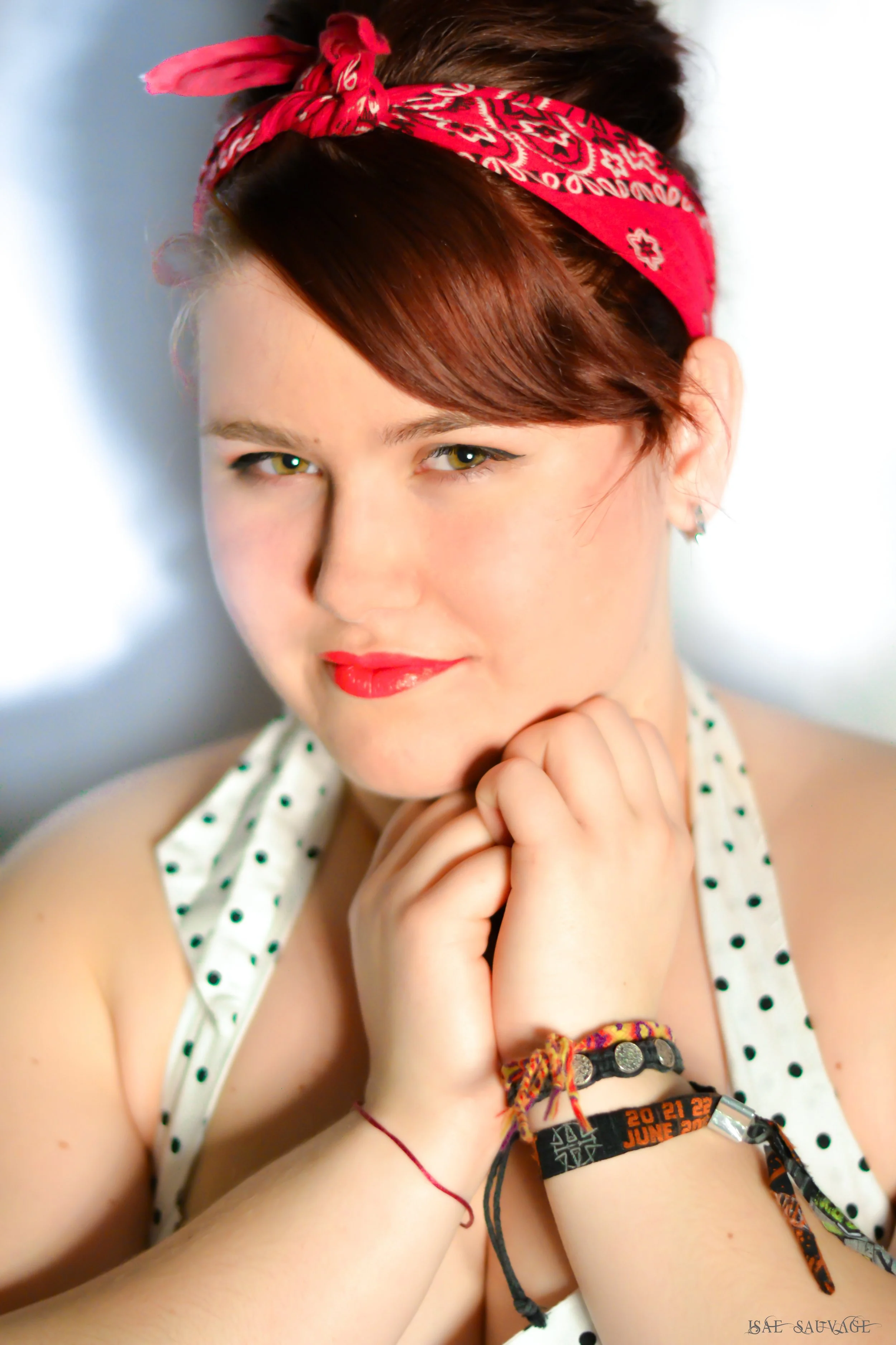 Une jeune femme avec des cheveux bruns, portant un bandana rouge, sourit légèrement, portant une blouse blanche à pois verts, avec plusieurs bracelets colorés et un bracelet à la montre sur le poignet droit, posant avec le menton posé sur ses mains. 