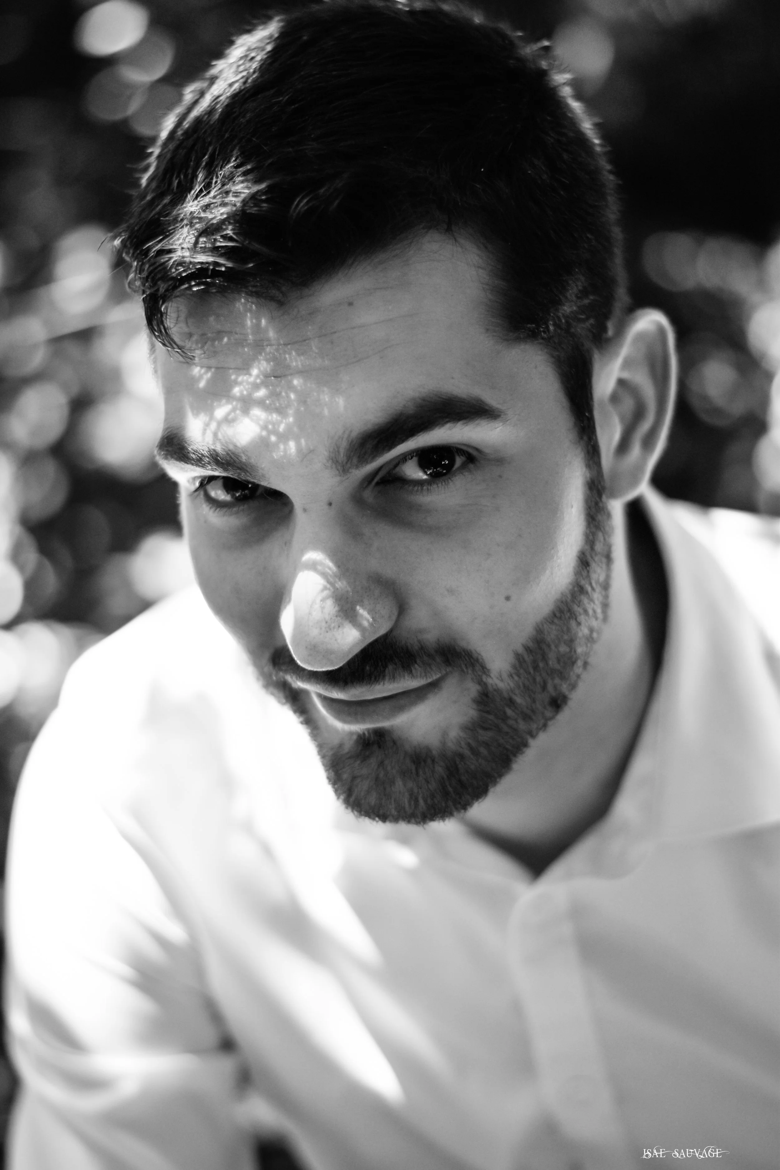 Un portrait en noir et blanc d'un jeune homme avec des cheveux courts, une barbe légère, regardant vers l'objectif, portant une chemise blanche, avec un fond flou d'éclats lumineux.