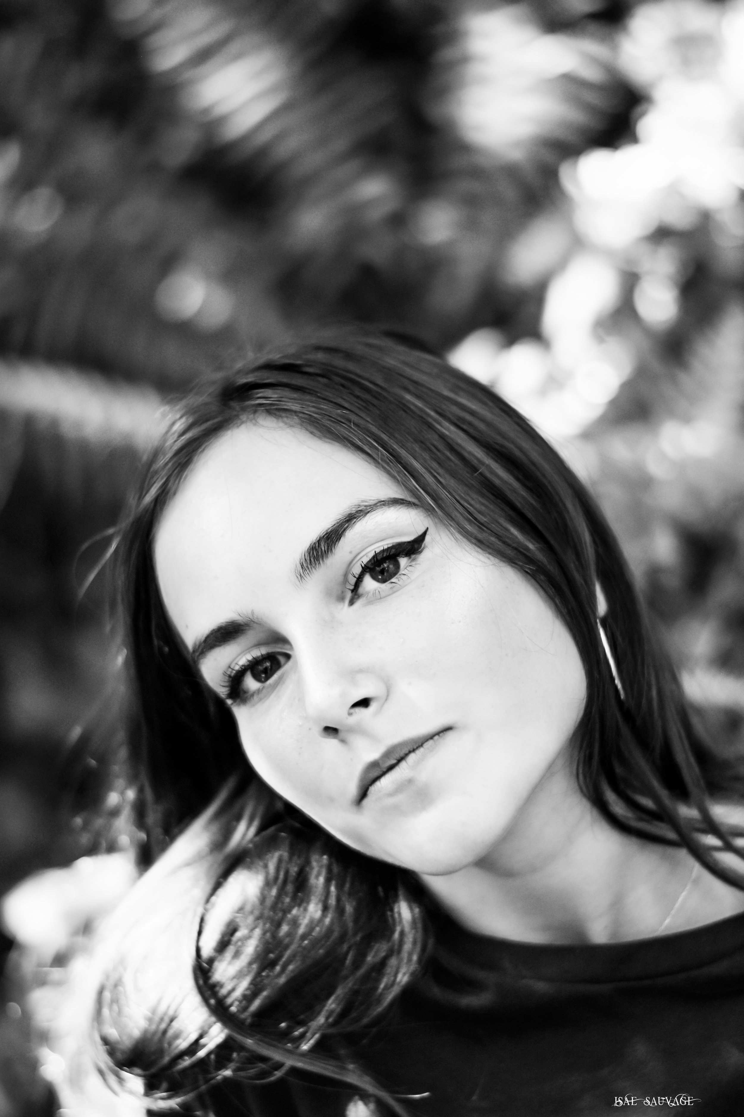 Un portrait en noir et blanc d'une femme avec des cheveux longs et foncés, ses yeux soulignés d'un maquillage noir, regardant vers la caméra. Fond avec des lumières d'arrière-plan.