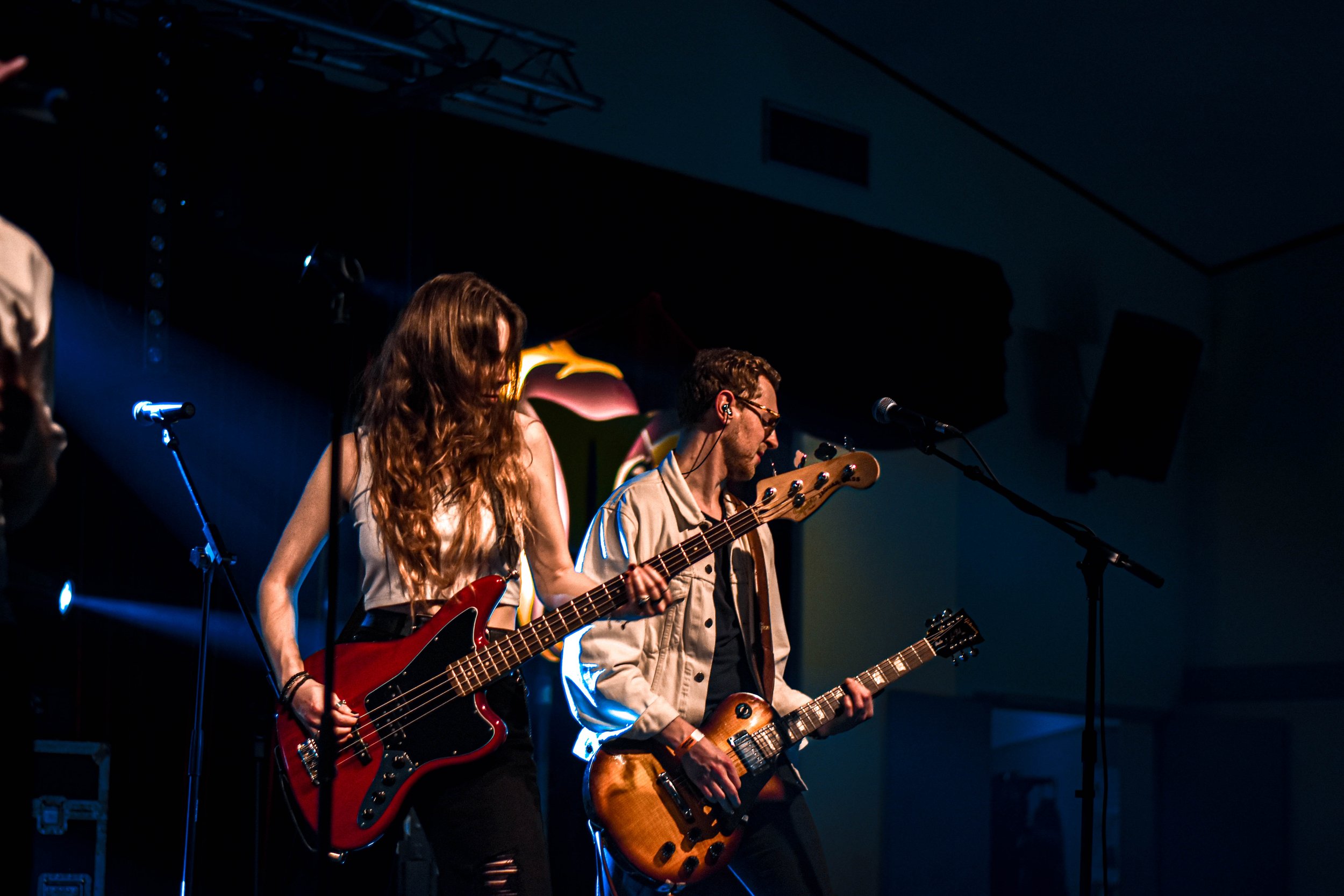 Deux musiciens jouent de la guitare sur scène lors d'un concert, sous un éclairage sombre, avec un fond coloré.