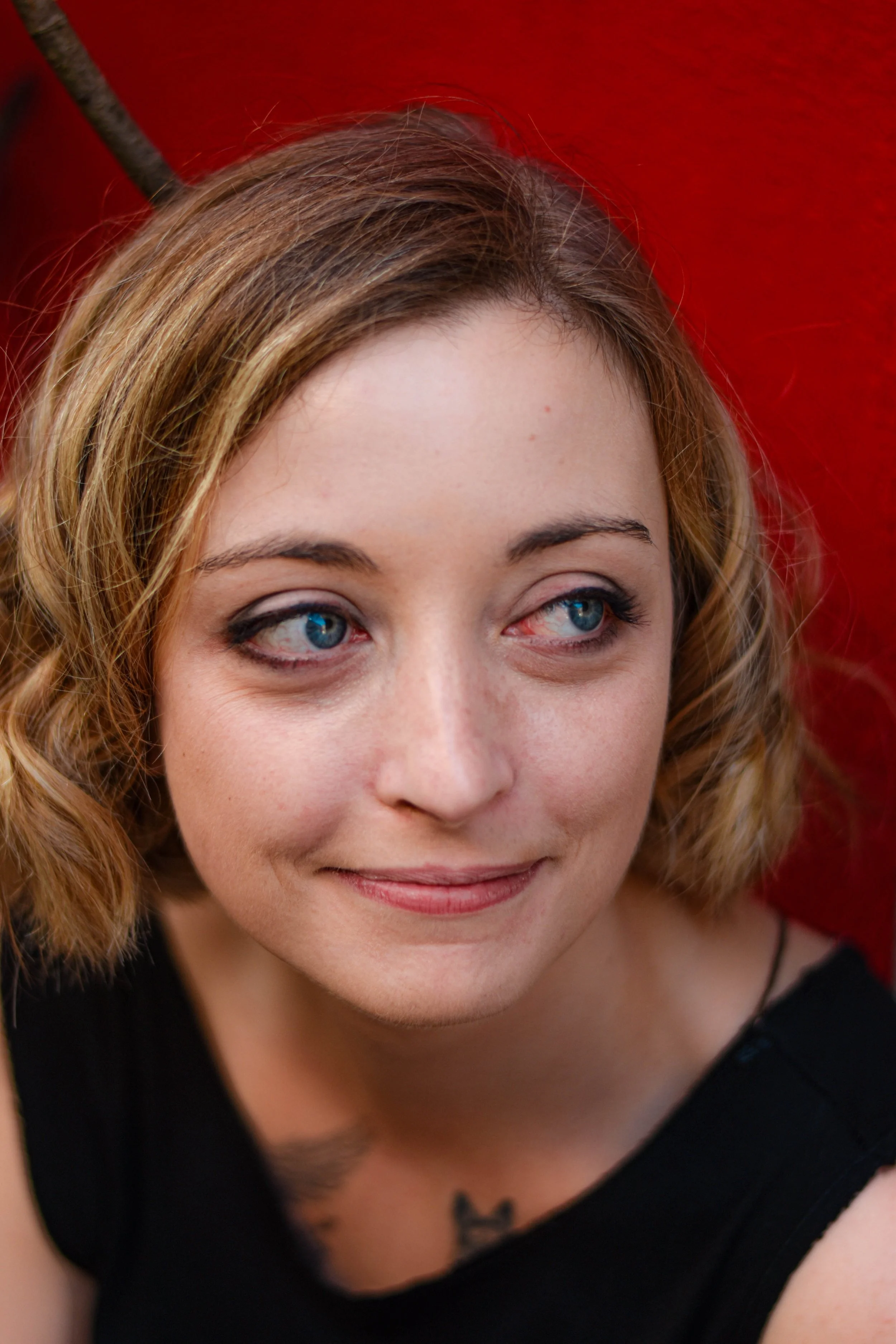 Une jeune femme aux cheveux bouclés, avec des yeux bleus, portant un haut noir, souriant légèrement, en arrière-plan rouge.