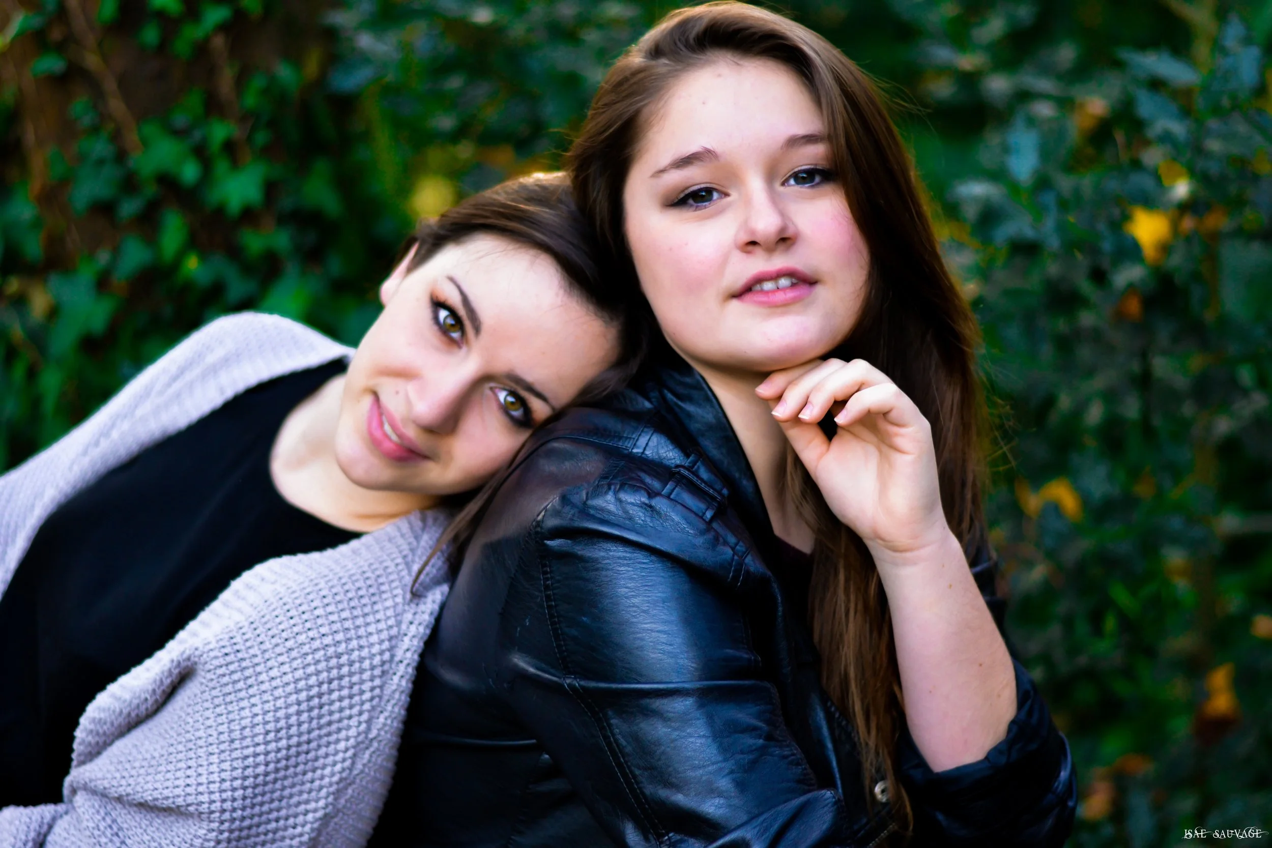 Deux jeunes femmes posent ensemble dans un environnement naturel avec des arbres verts en arrière-plan, l'une repose la tête sur l'épaule de l'autre.