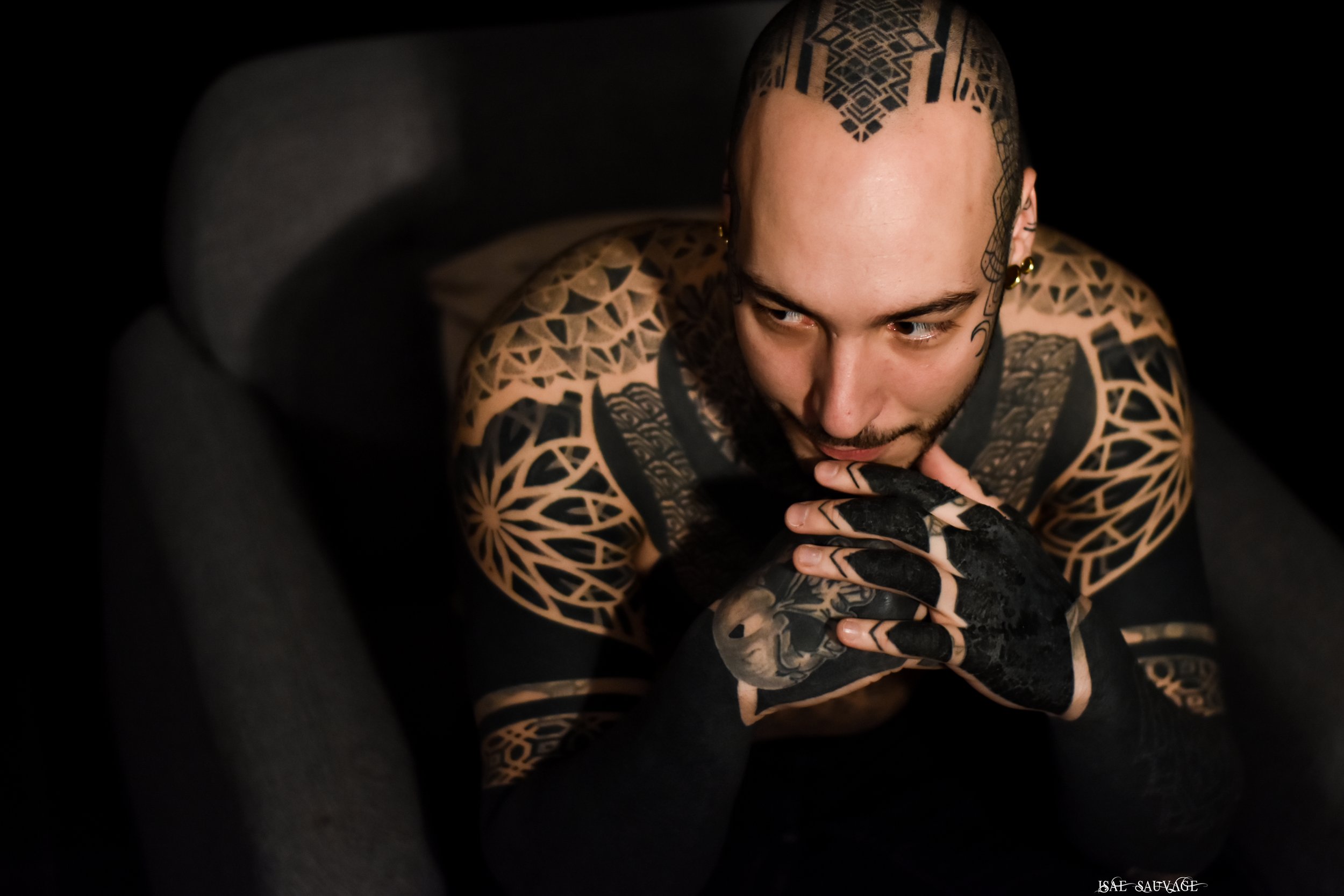Jeune homme avec tatouages couvrant la tête, les bras et les mains, posant en regardant légèrement vers le bas, dans un cadre sombre.