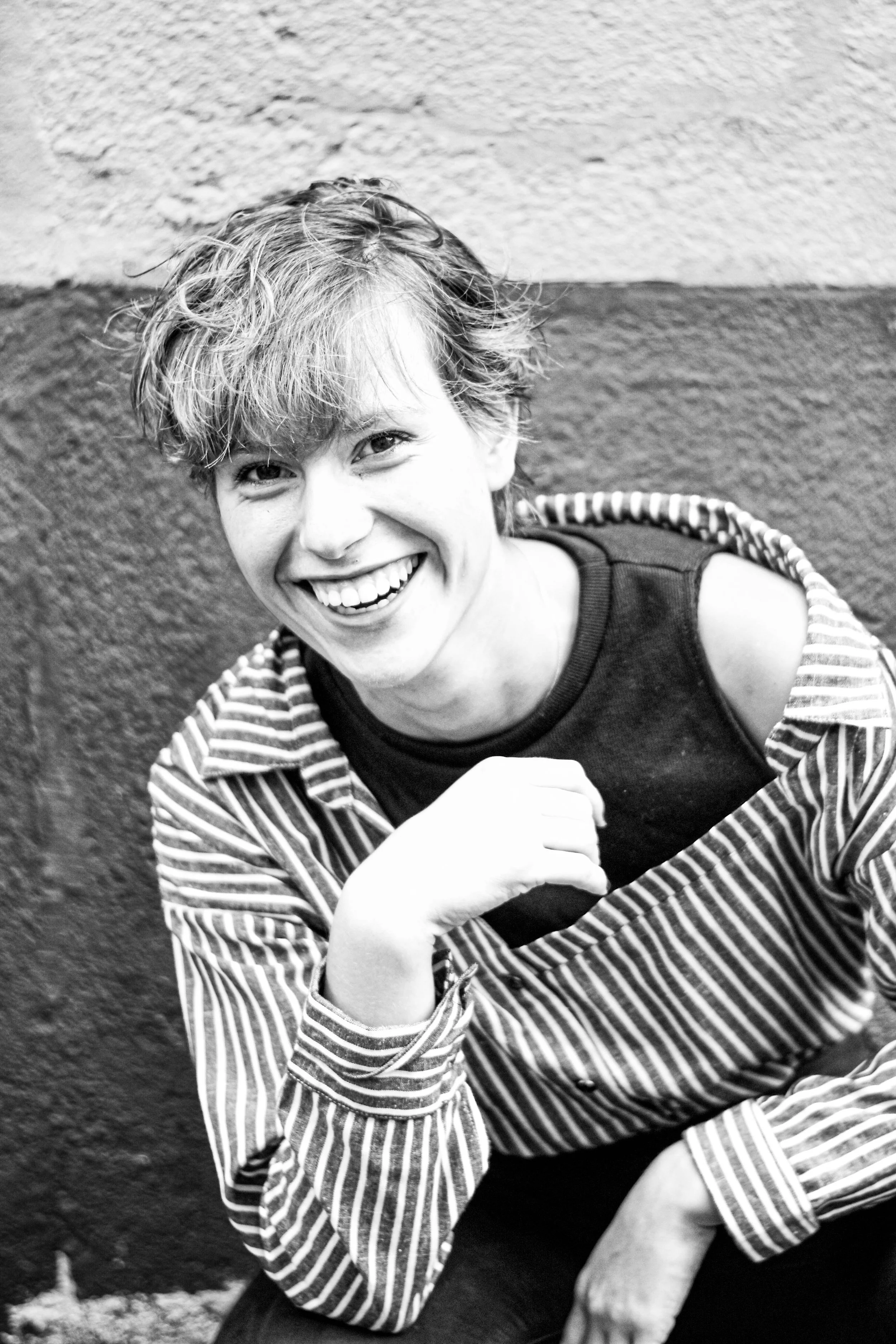 Une femme souriante avec des cheveux courts et bouclés, portant un t-shirt noir et une veste à rayures blanches et noires, assise contre un mur texturée, en noir et blanc.
