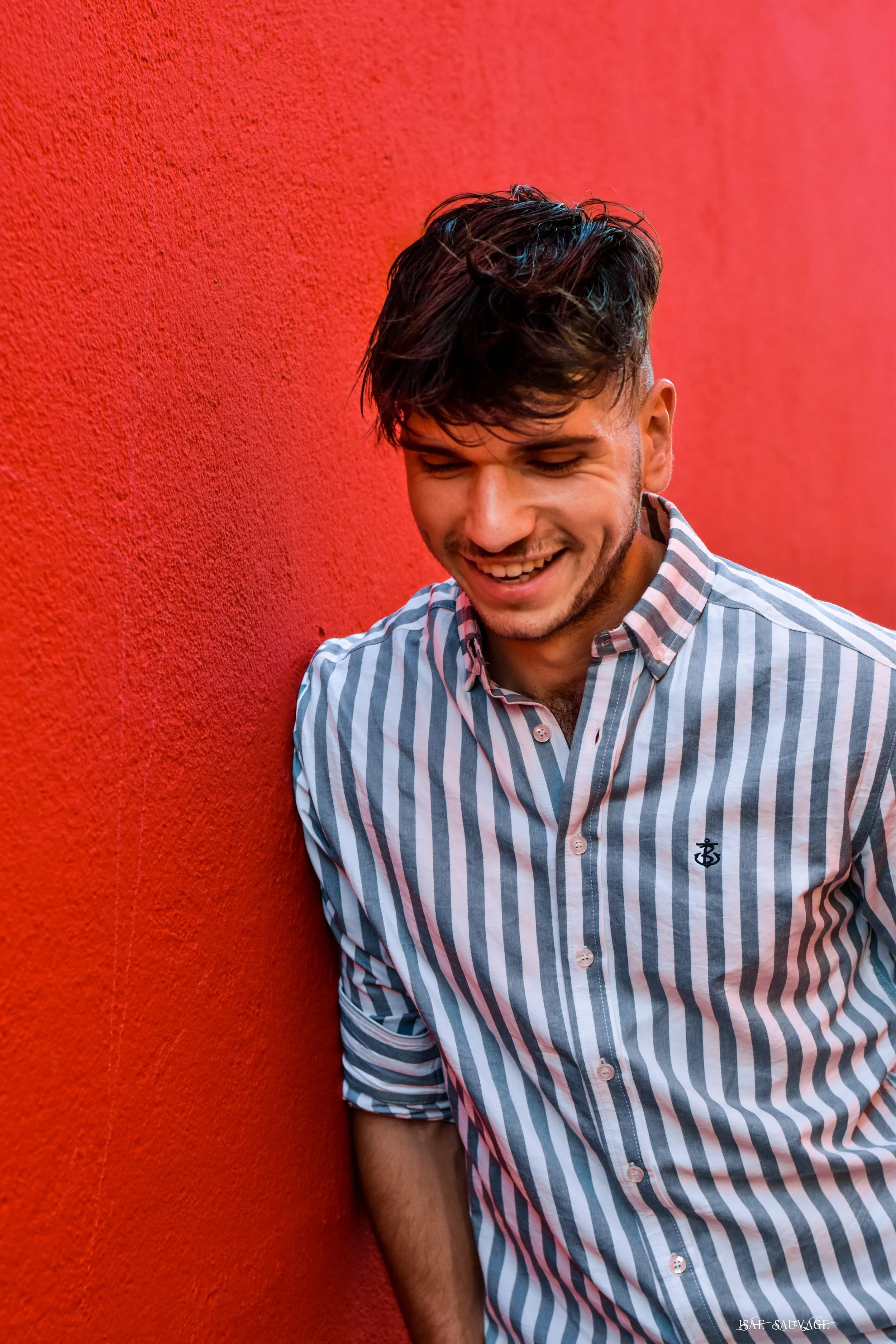 Jeune homme souriant portant une chemise à rayures claires et foncées, debout contre un mur rouge.