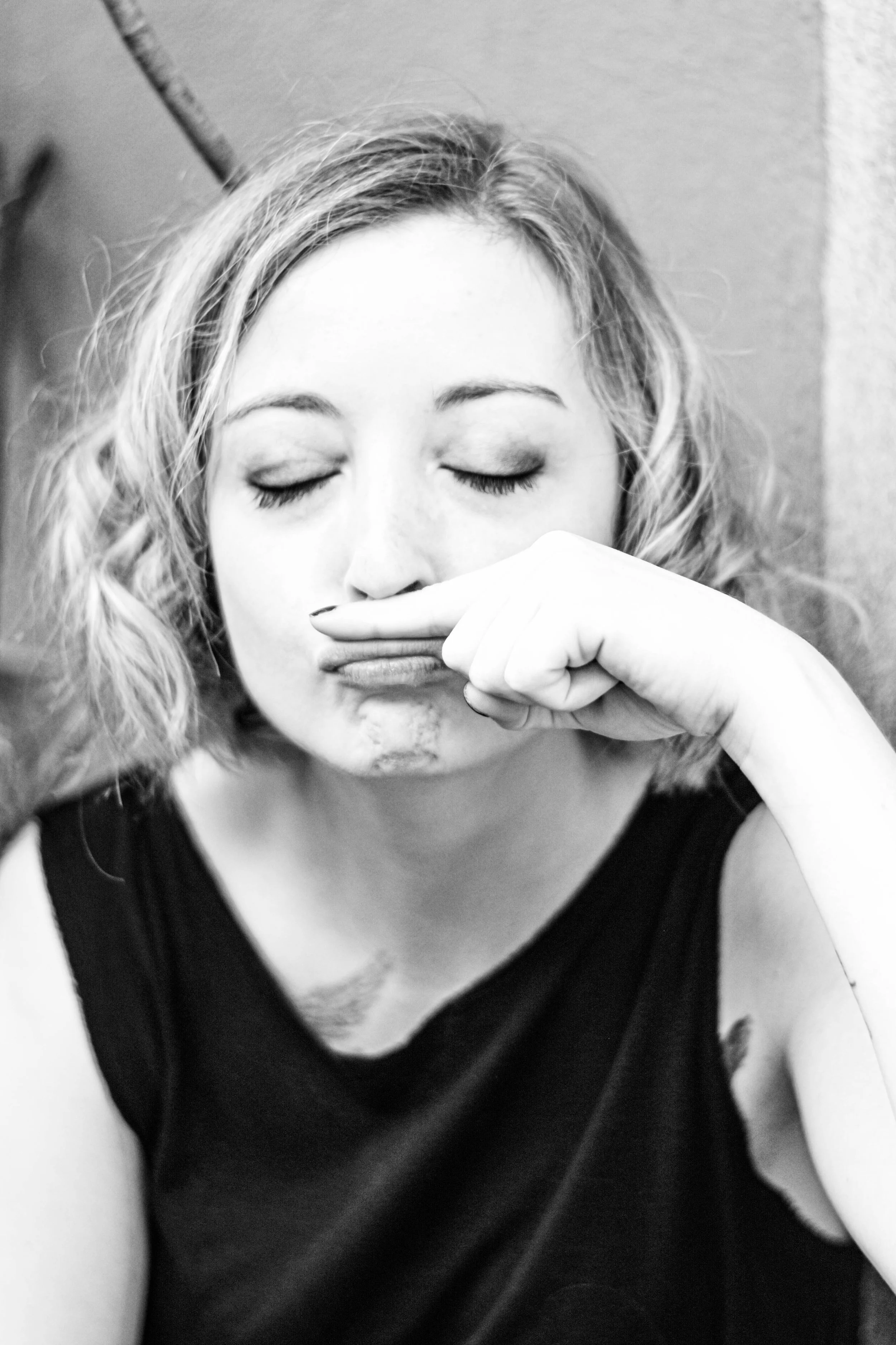 Une femme avec des cheveux bouclés qui fait une grimace avec sa main, portant un haut noir, assise contre un mur, en noir et blanc.