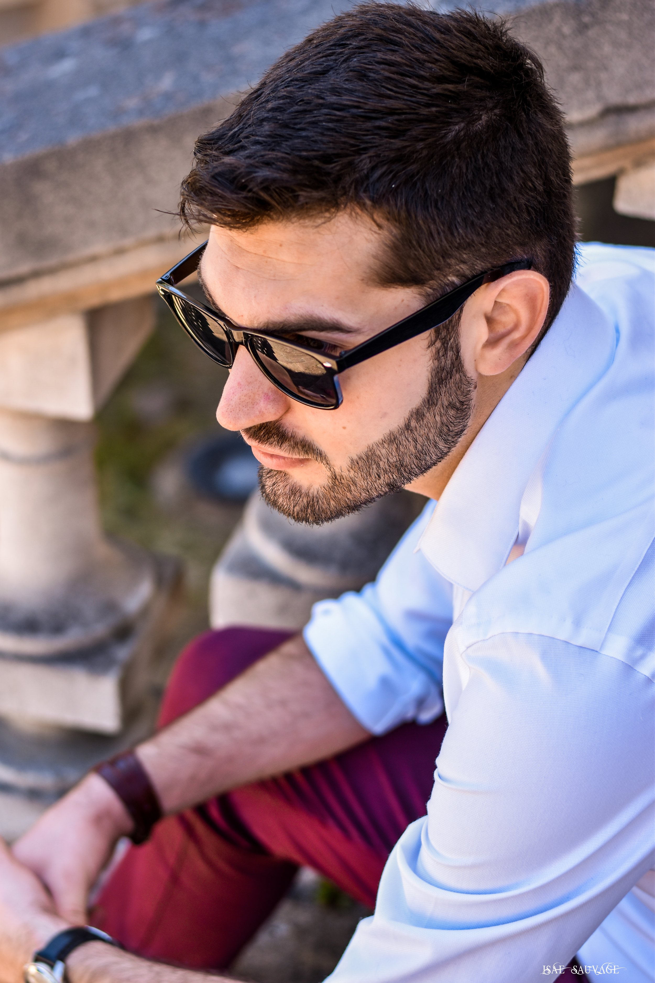 Un homme portant des lunettes de soleil et une chemise blanche regarde vers le bas, à l'extérieur, avec des bancs en pierre en arrière-plan.
