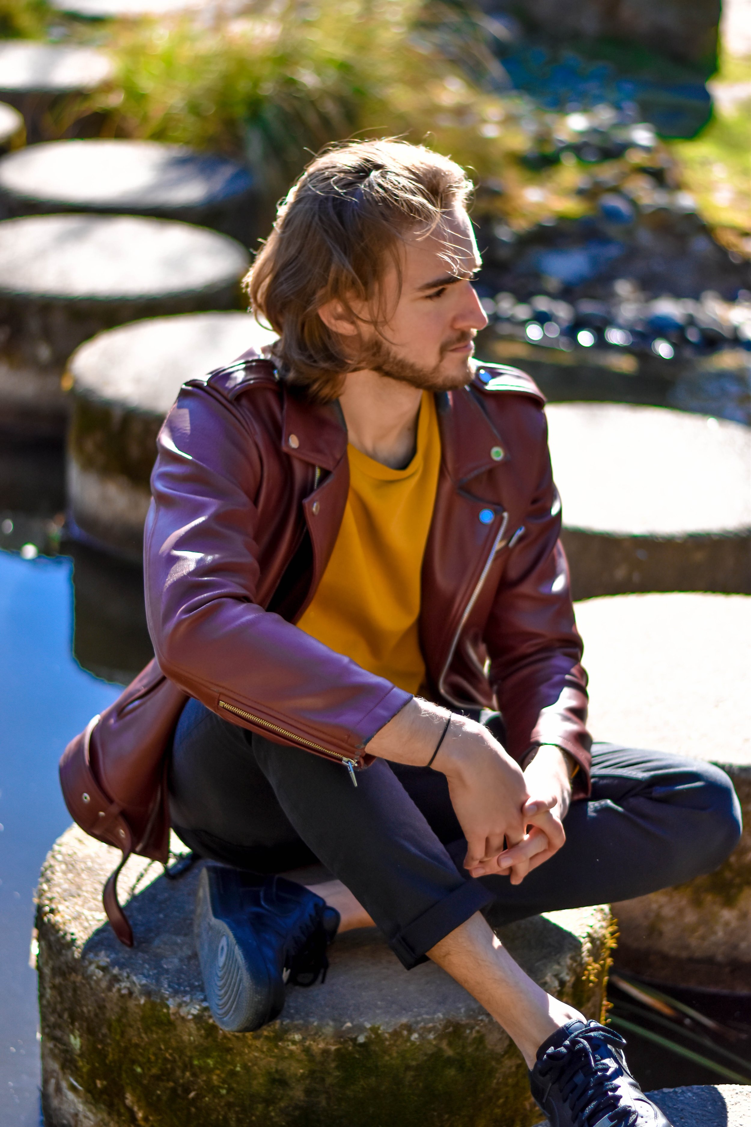 Un jeune homme avec des cheveux longs, portant une veste en cuir marron, un t-shirt jaune et des baskets noires, assis sur un rocher près d'un plan d'eau à l'extérieur.