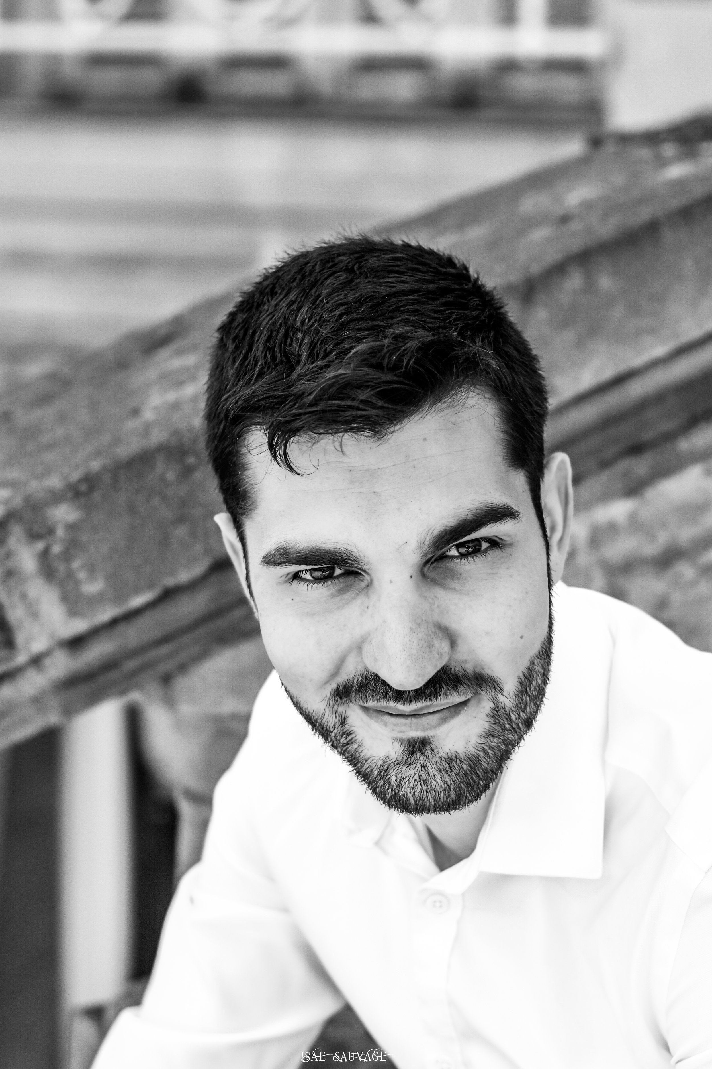 Portrait en noir et blanc d'un homme avec une barbe et des cheveux foncés courts, portant une chemise blanche. Le fond est flou.