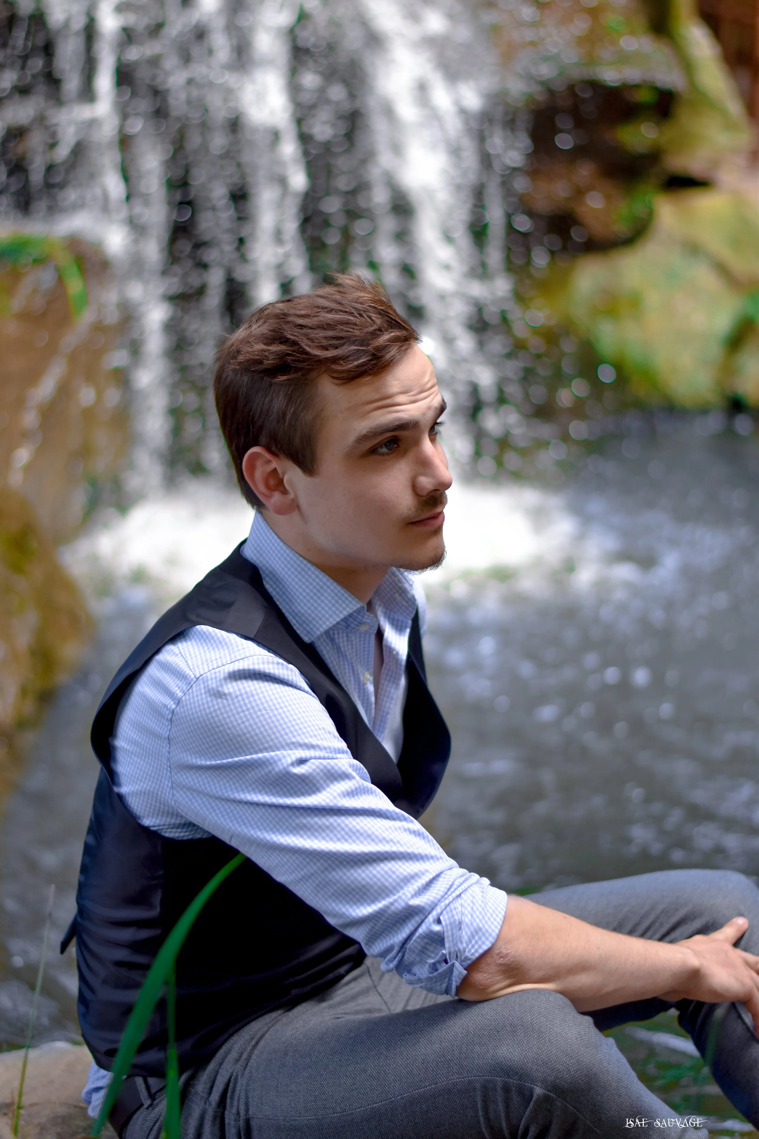 Jeune homme assis près d'une cascade, portant une veste noire, une chemise à carreaux bleus et une cravate, avec des cheveux bruns, en pleine nature.